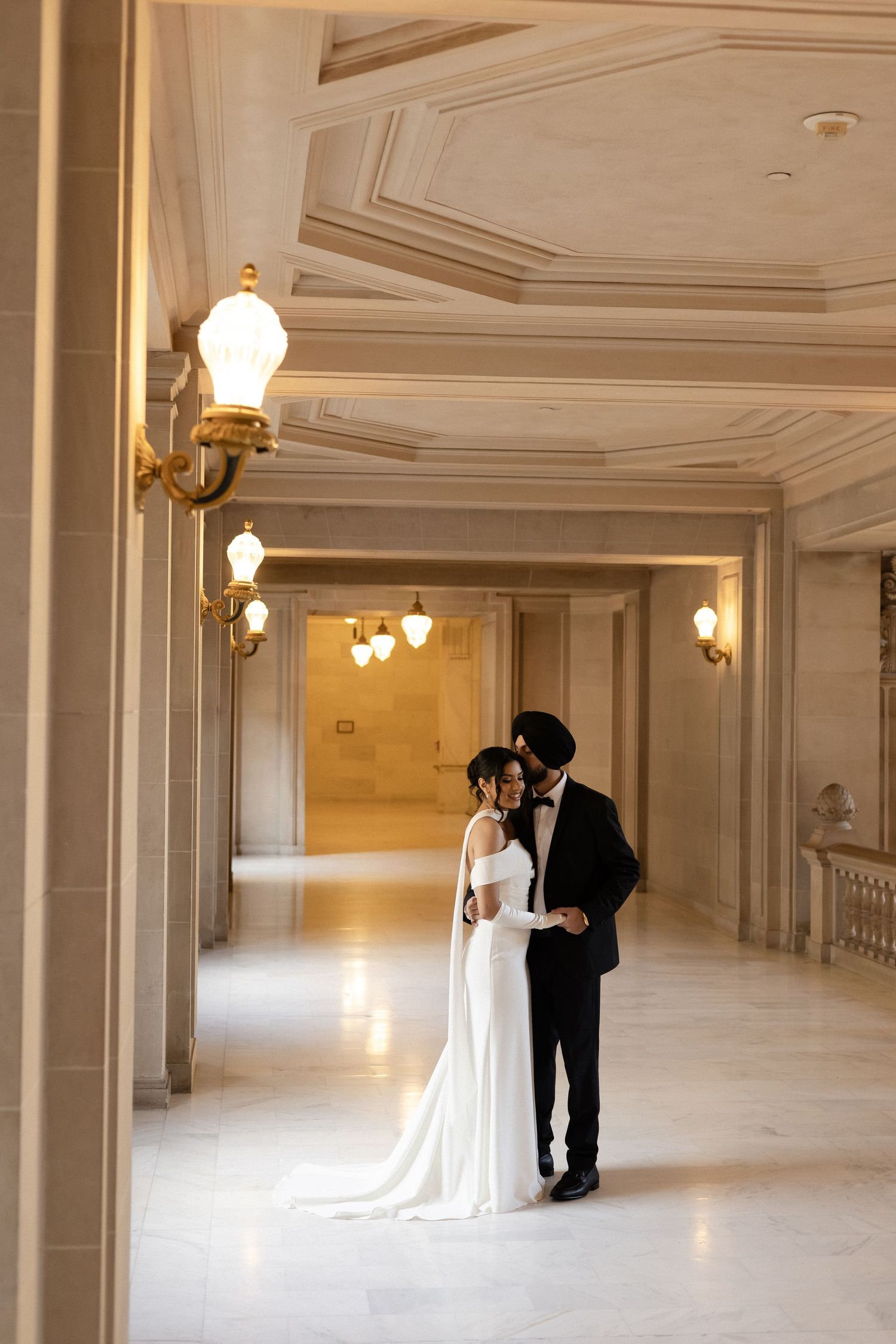 Elegant couple in formal wedding attire stands in ornate marble hallway with classical architectural details and wall sconces.
