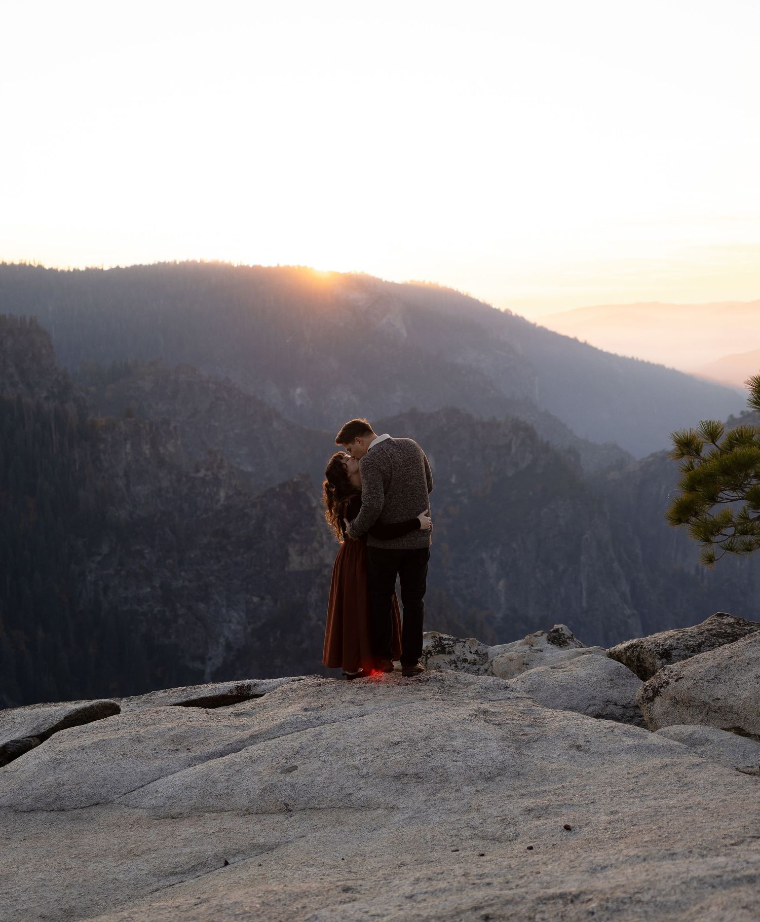 Two people sit together on a rocky mountain ledge watching the sunset.