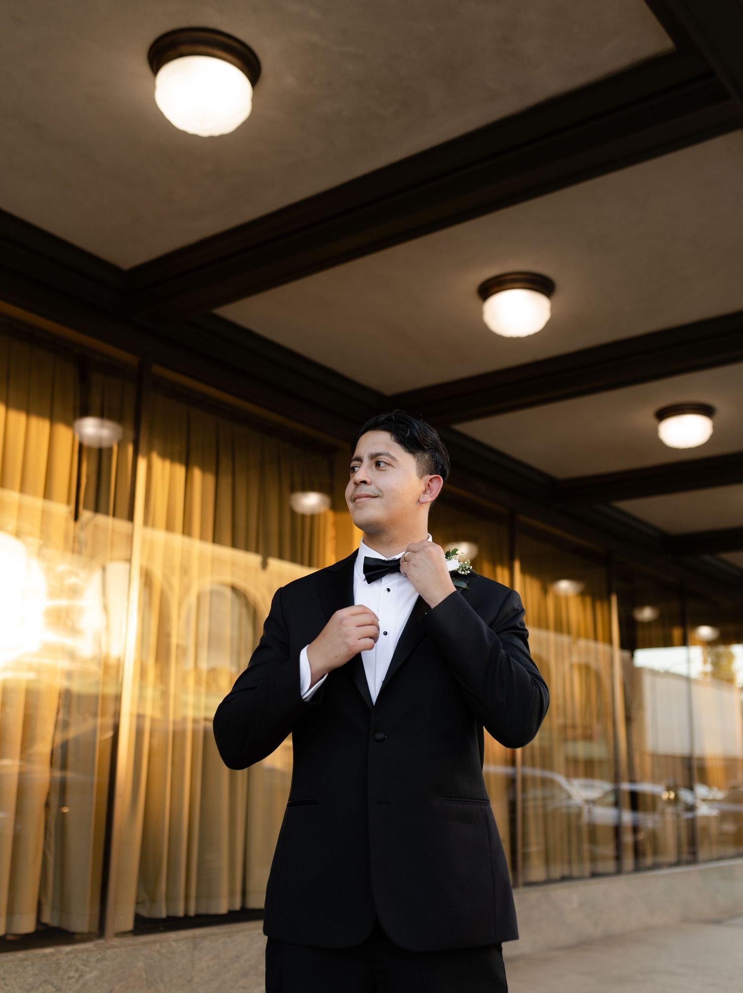 Person in formal black attire adjusts their bowtie in a warmly lit indoor setting.