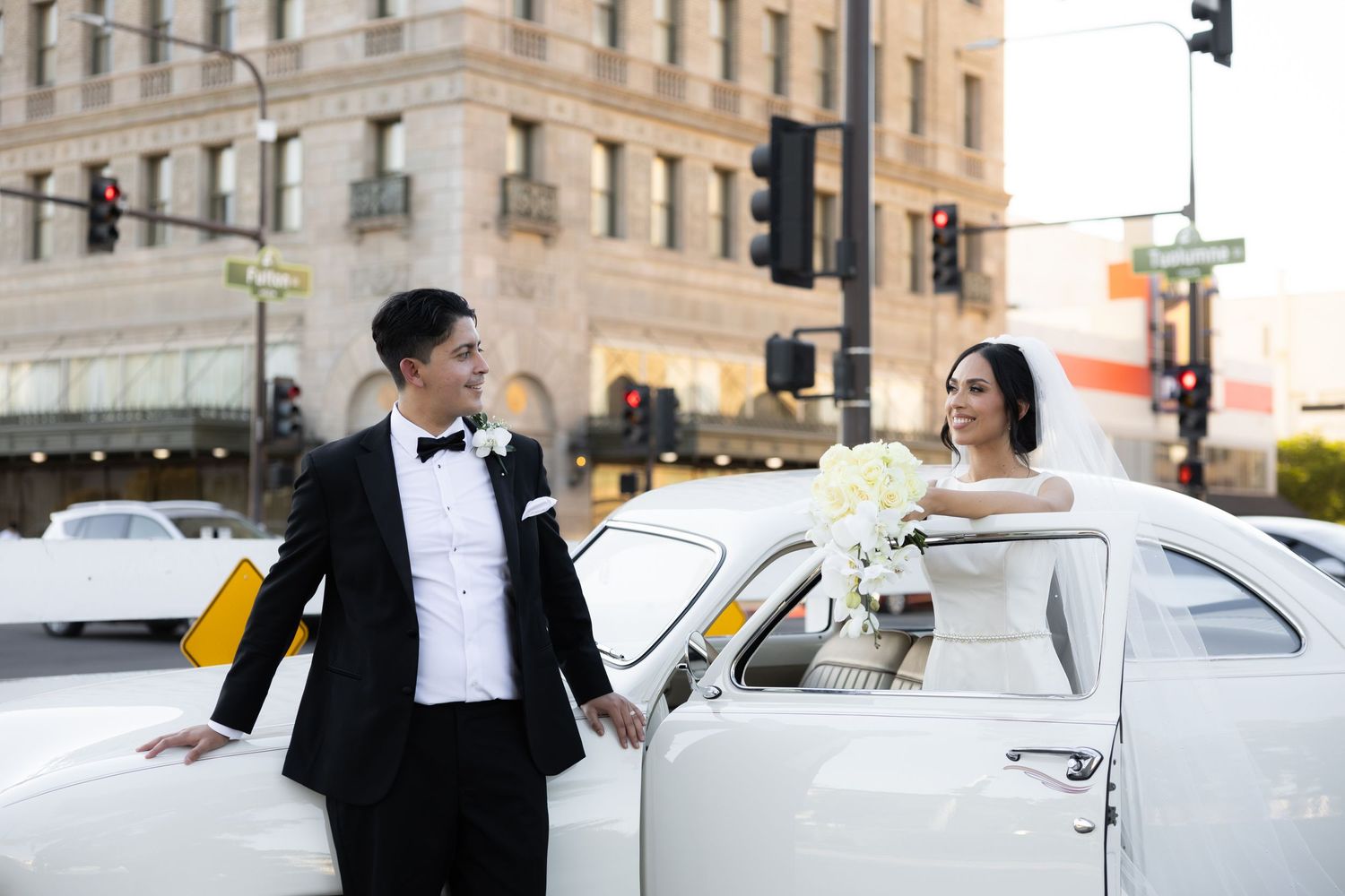 Couple in wedding attire poses with vintage white car on city street.