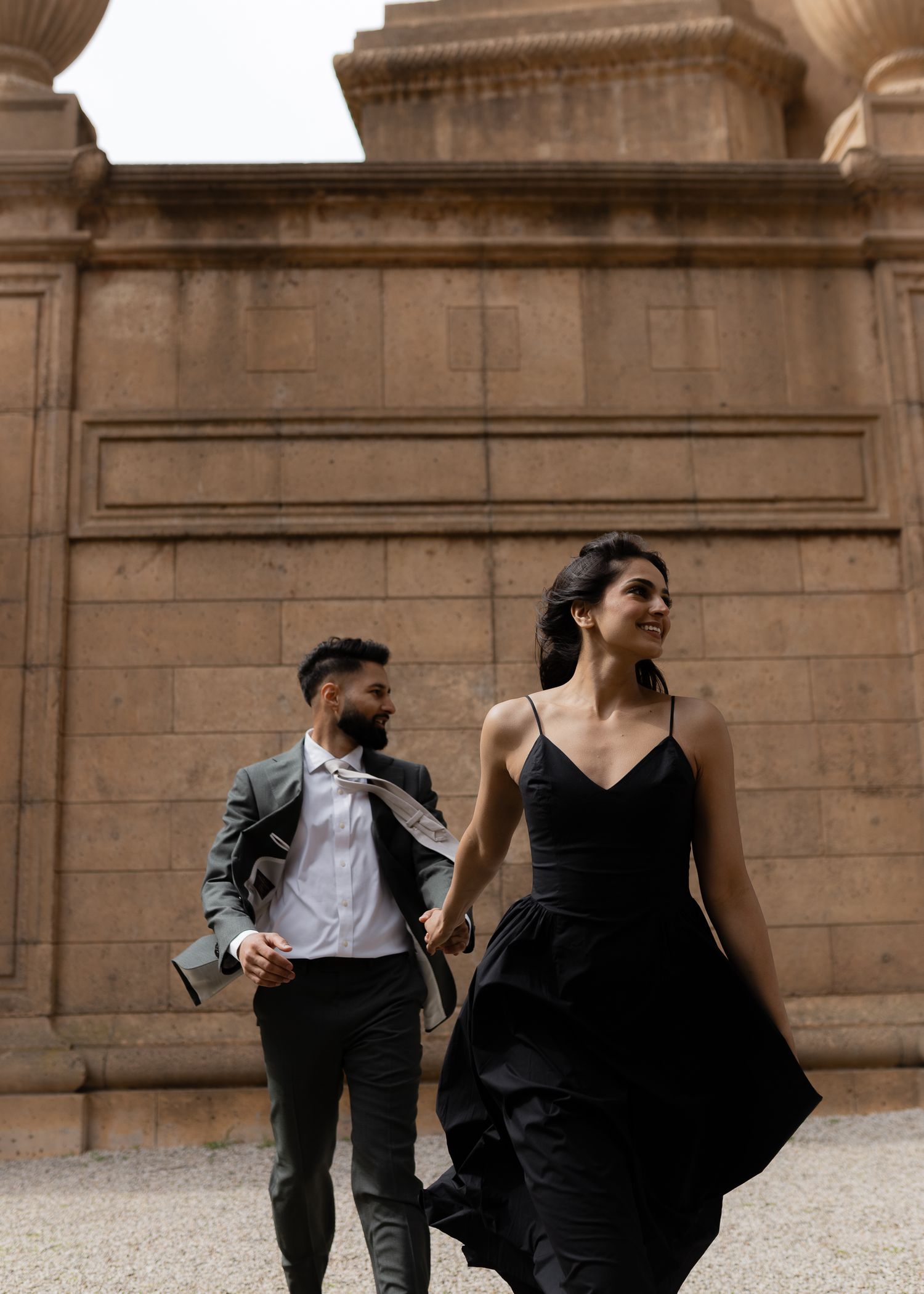 A couple in formal attire poses against stone architecture with flowing black dress.