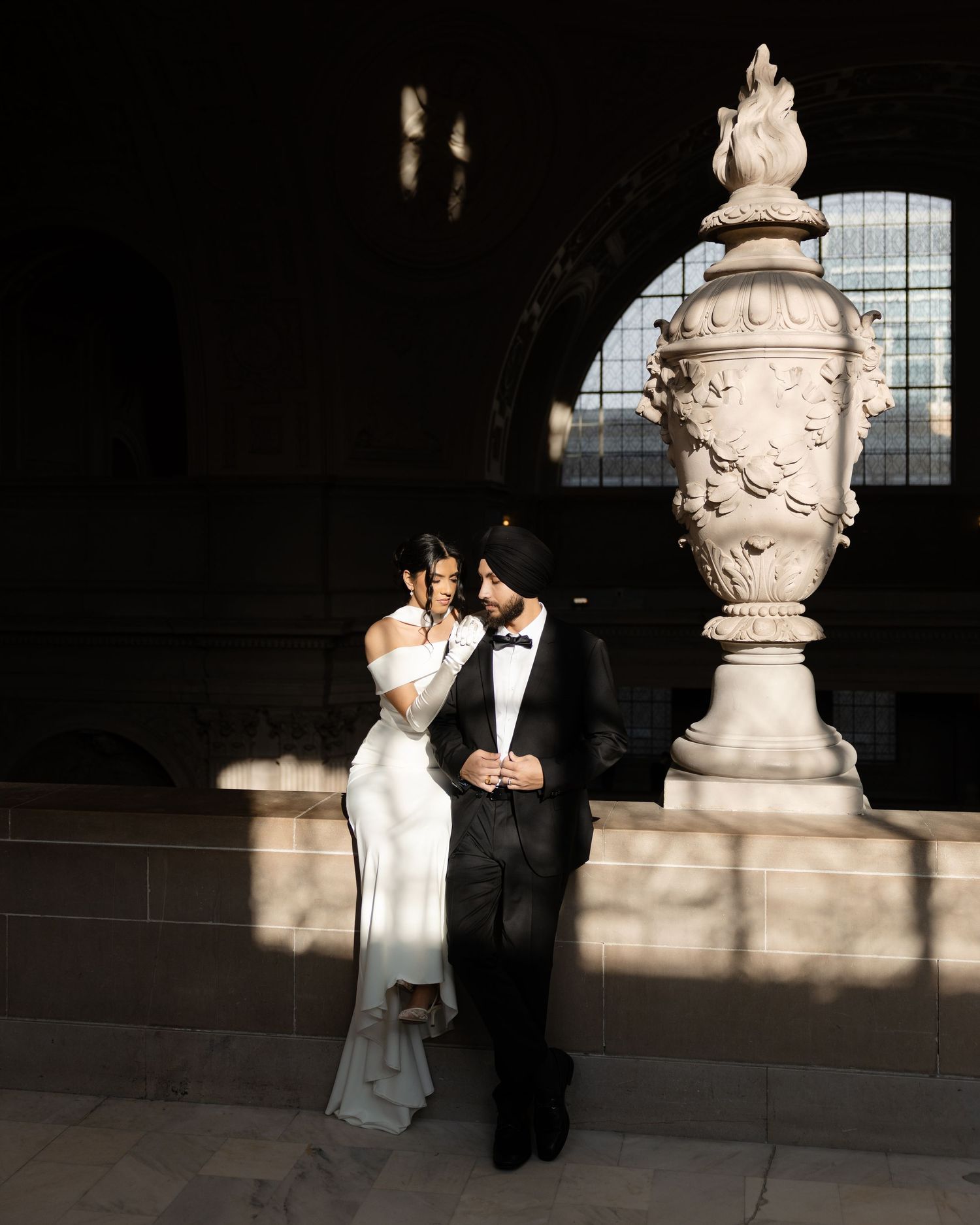 A couple poses in formal attire against a dark background with ornate architectural details.