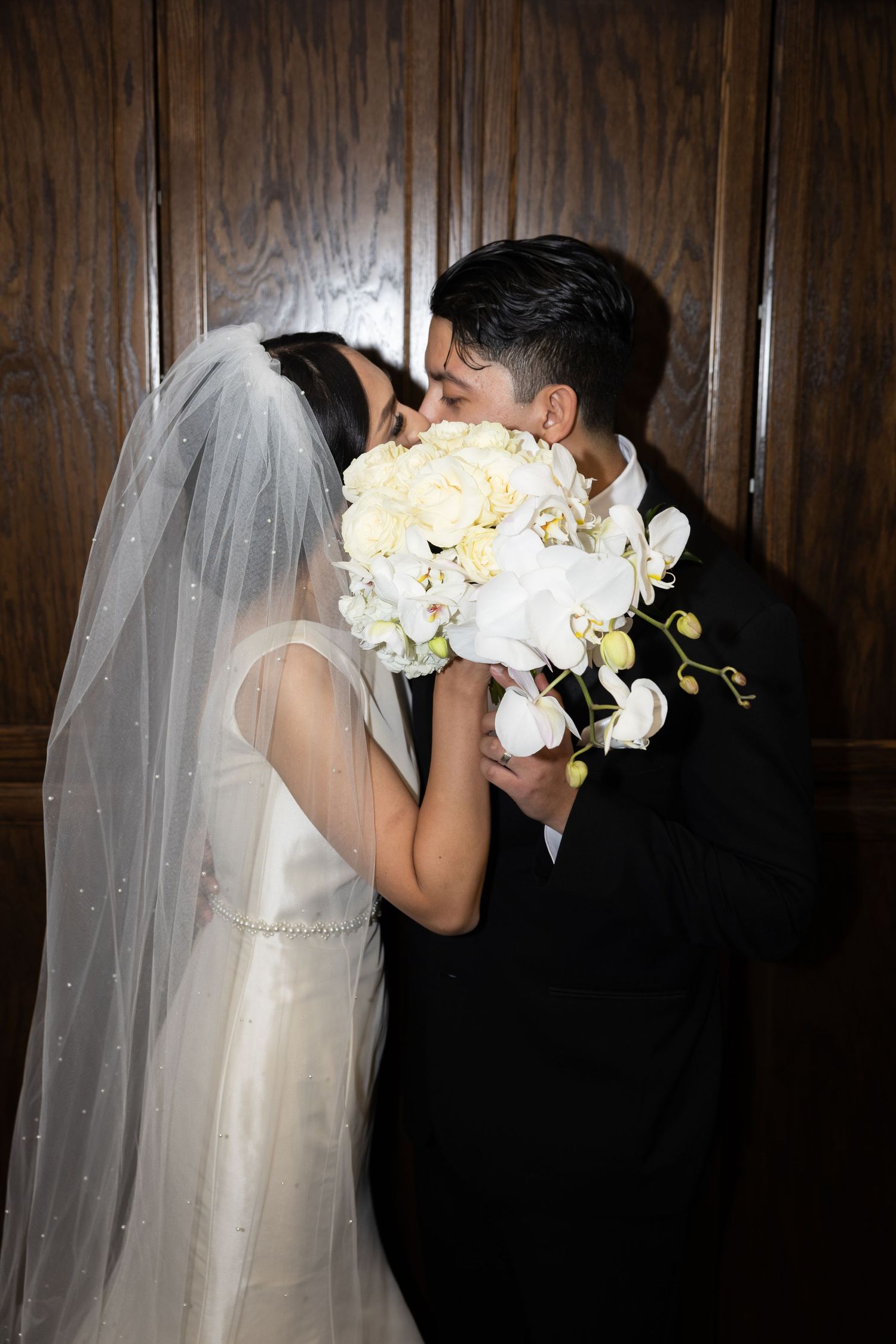 Romantic wedding moment captured against dark wood paneling with white floral bouquet.