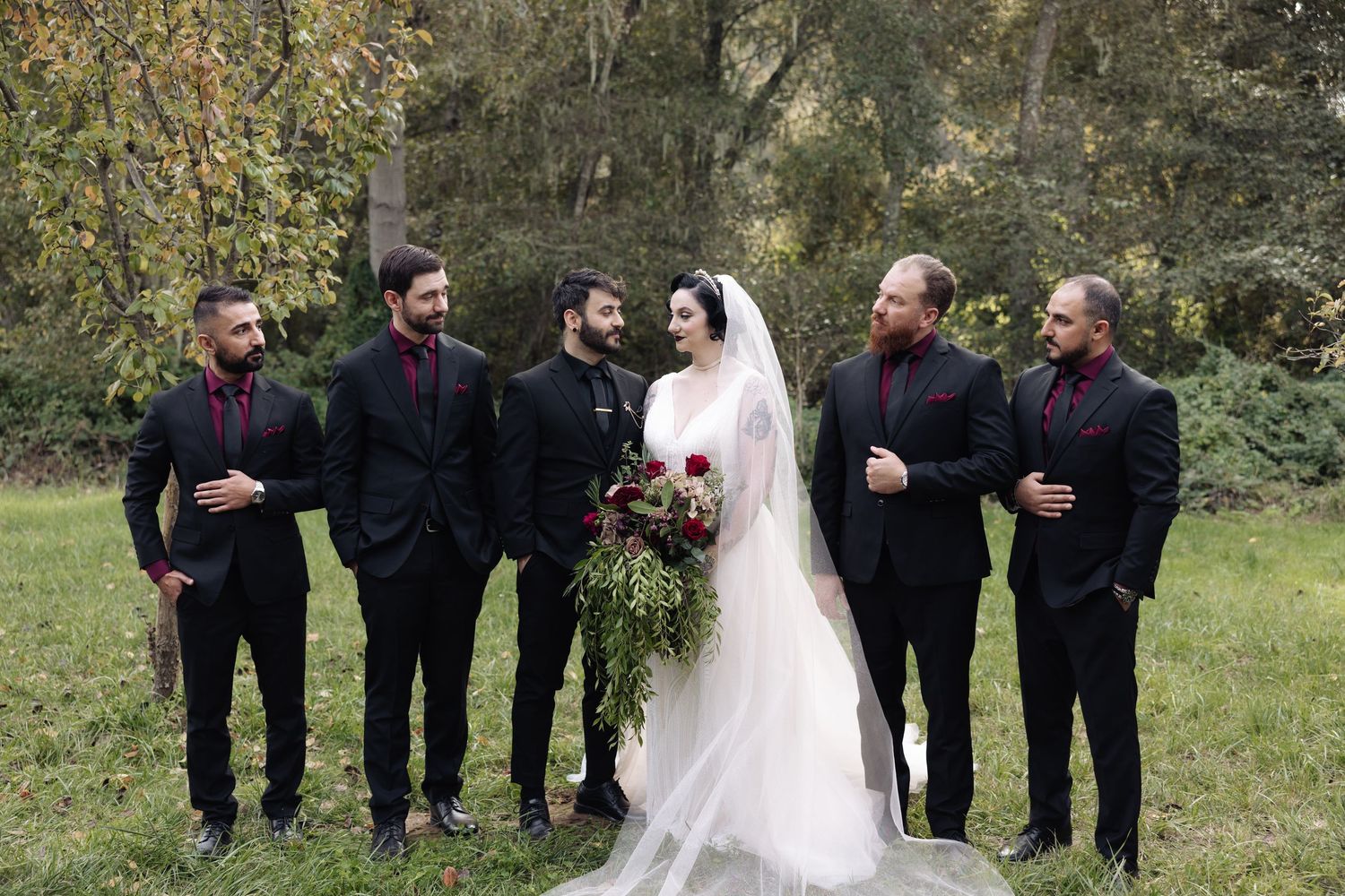 Wedding party stands in a line outdoors with groomsmen in black and bridesmaids in burgundy.