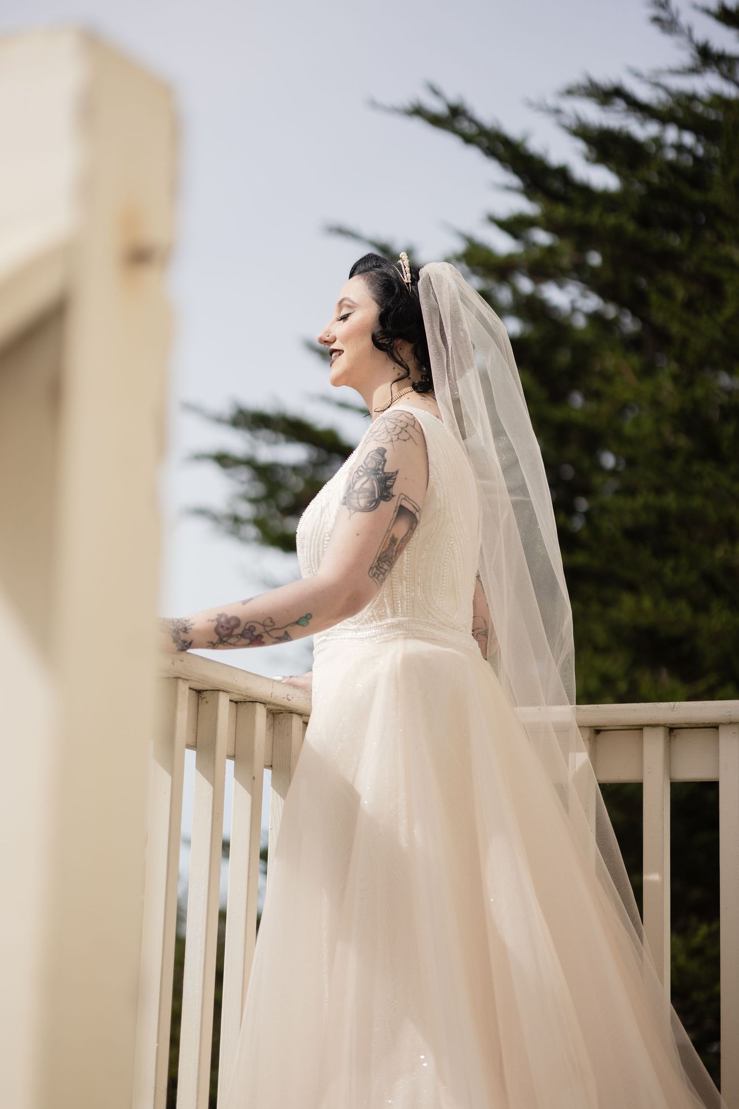Bride in flowing white dress and veil standing on outdoor deck with joyful expression.