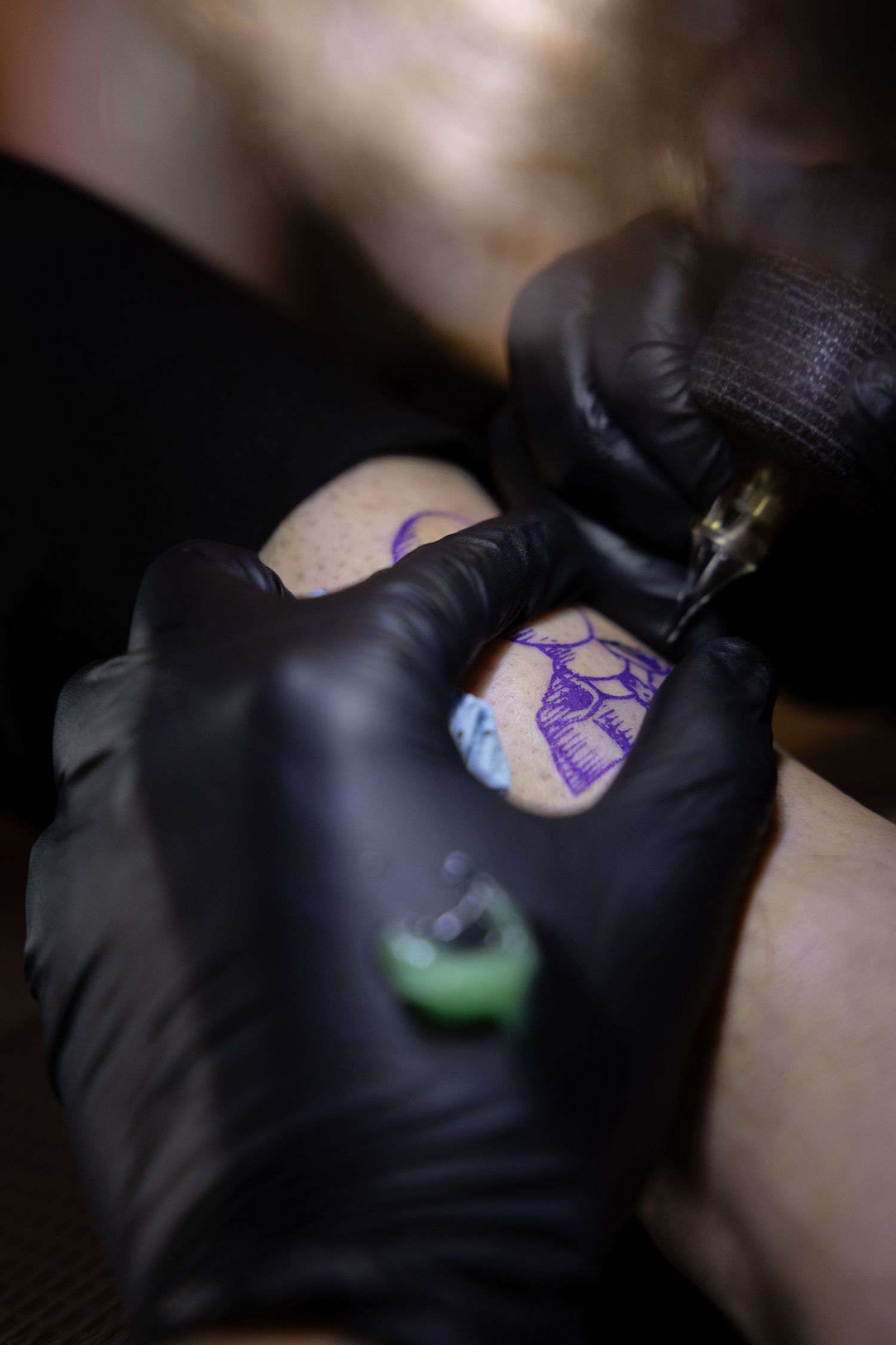 Close-up of a tattoo artist wearing black gloves working on a purple tattoo design.