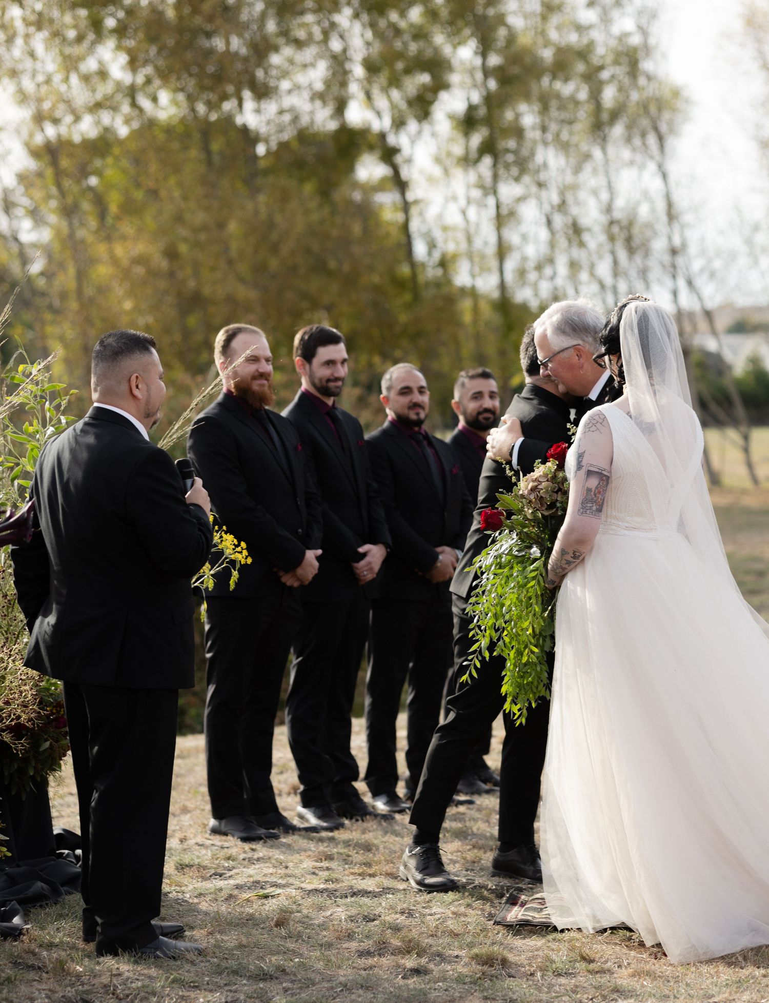 Wedding ceremony processional with groomsmen in black suits lined up outdoors.