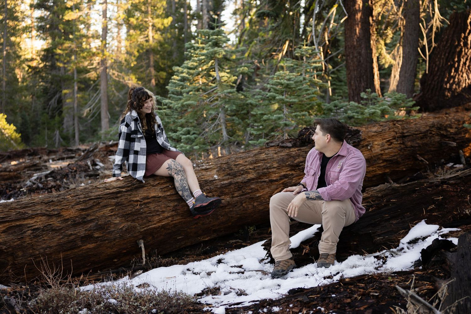 Couple sharing a romantic moment on a fallen tree trunk in a snowy forest setting.