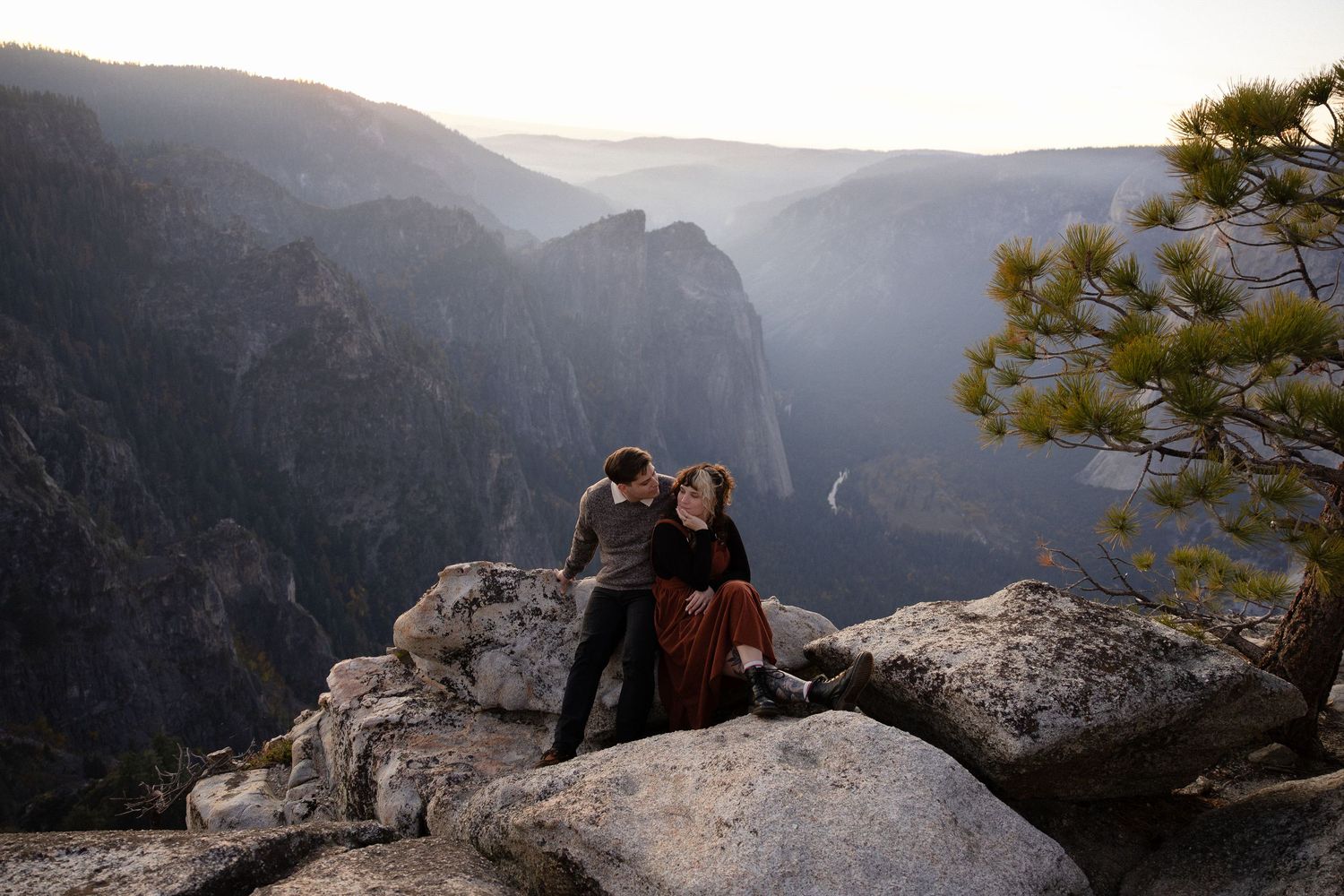 Two people embrace while laying on rocky mountain ledge with scenic valley backdrop.