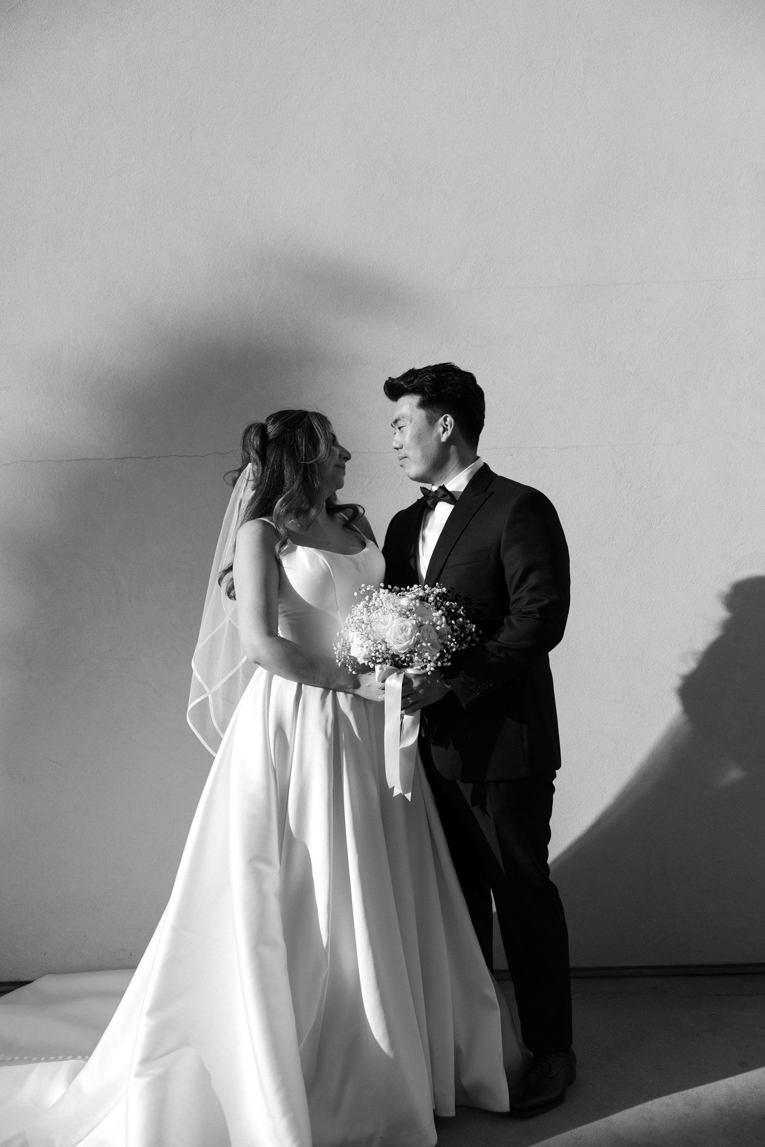 Black and white wedding photo of bride in white gown with veil and groom in tuxedo holding bouquet, gazing at each other.