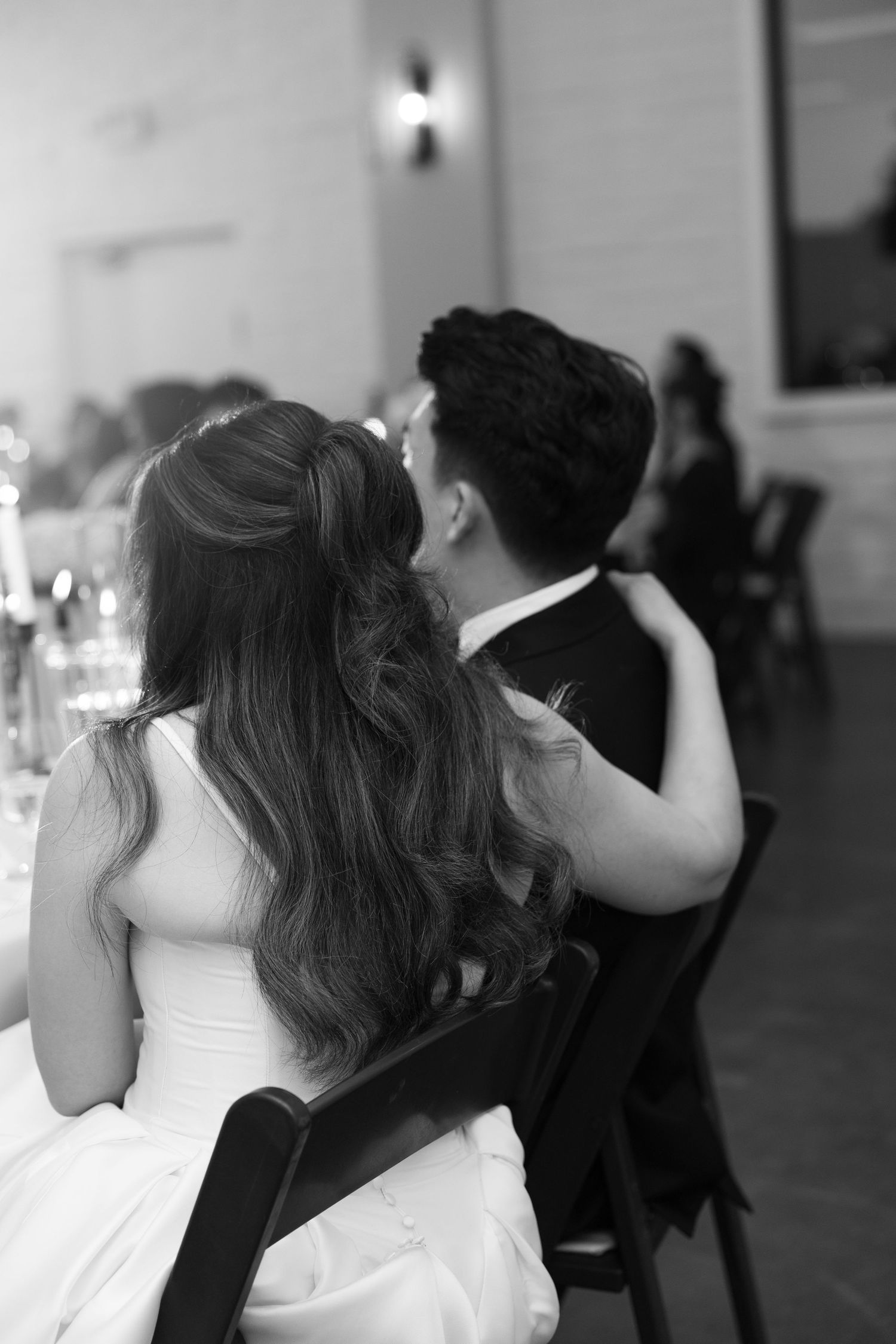 Black and white photo of bride and groom seated together at wedding reception, viewed from behind.
