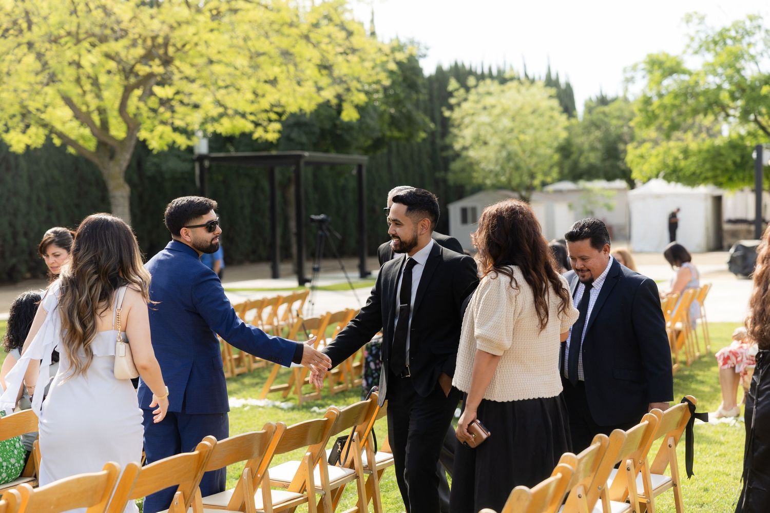 Guests greeting each other with handshakes at an outdoor wedding ceremony with wooden chairs on a sunny day.