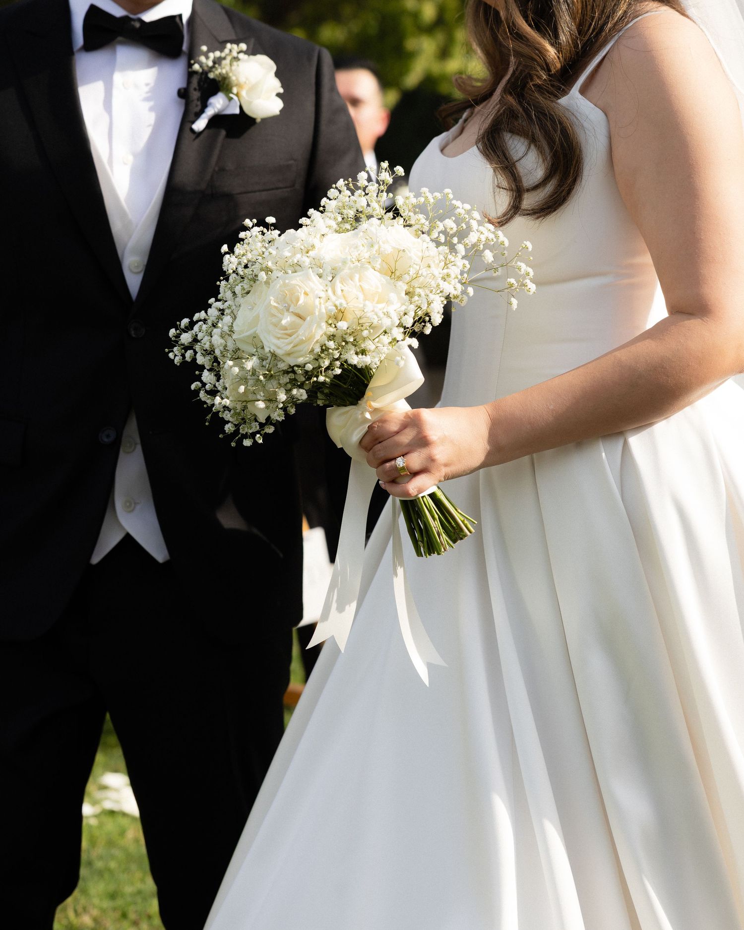 Close-up of bride holding white rose and baby's breath bouquet beside groom in tuxedo.