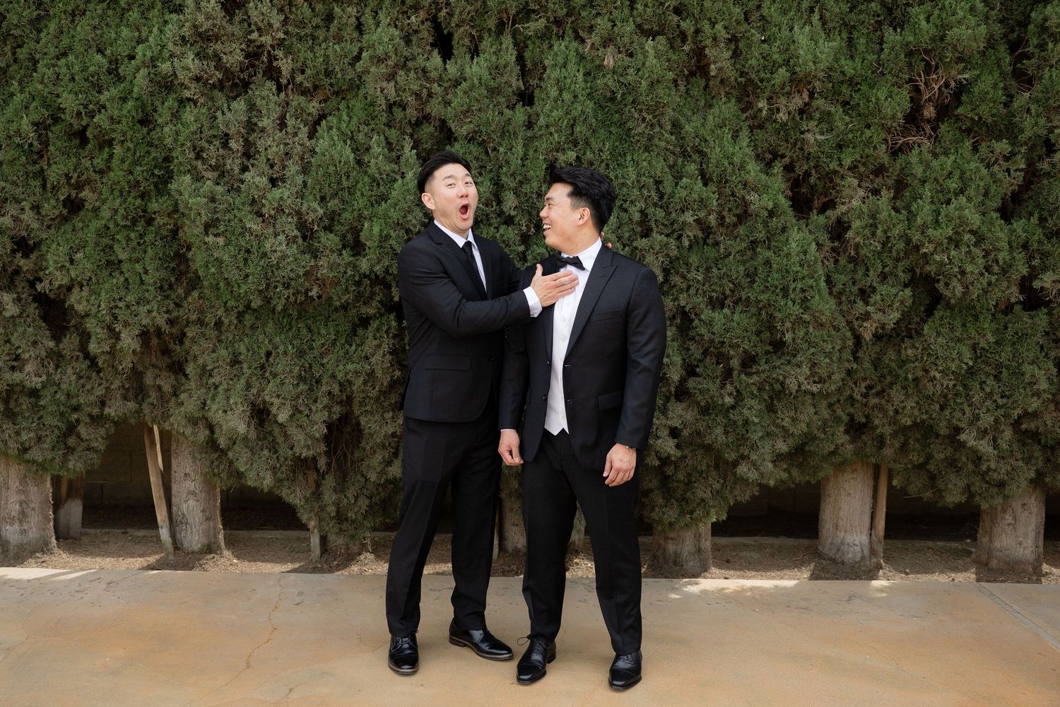 Two men in black tuxedos laughing together in front of green hedges at a wedding celebration.