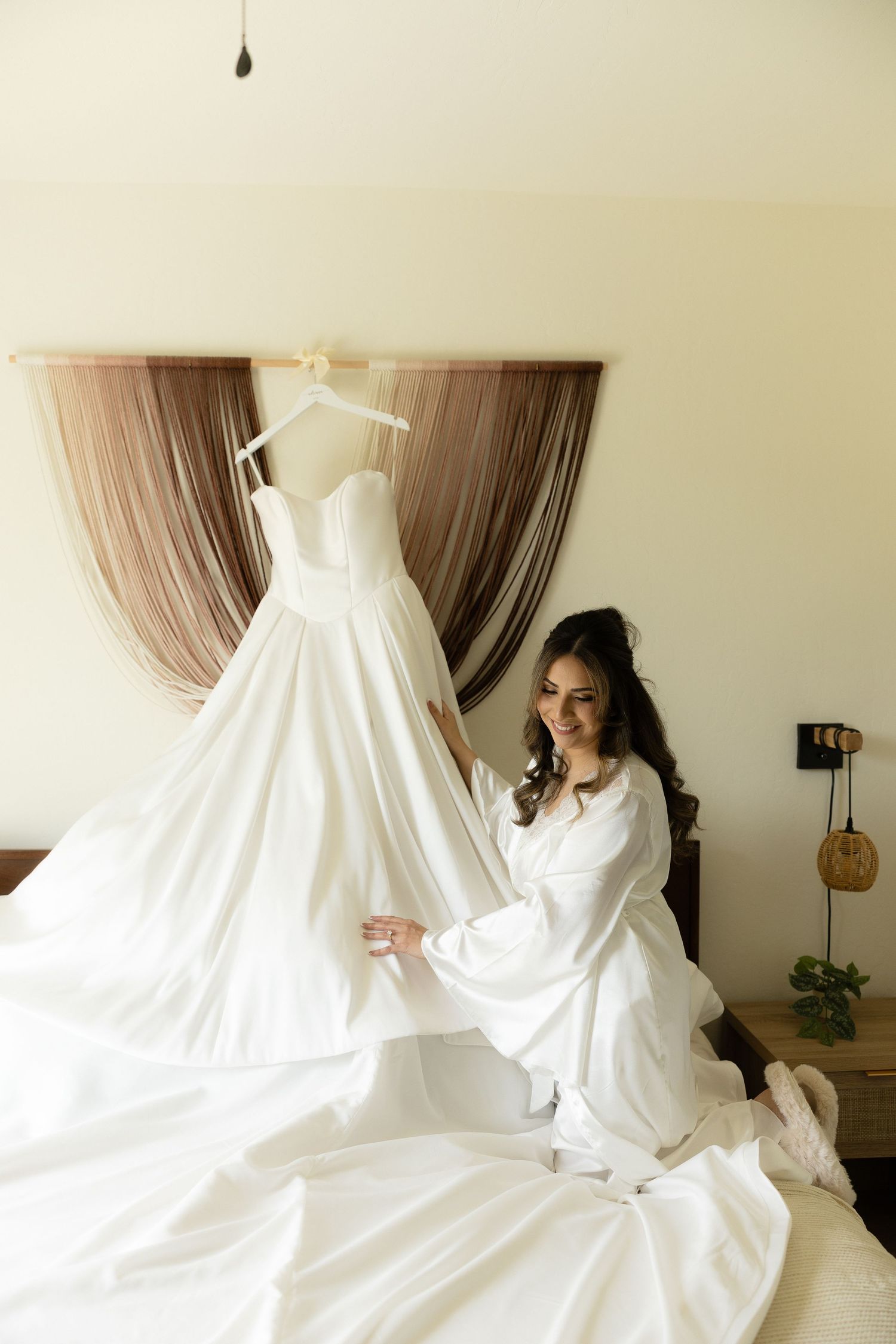 Smiling bride in white robe sitting on bed admiring her elegant white wedding gown on a hanger.