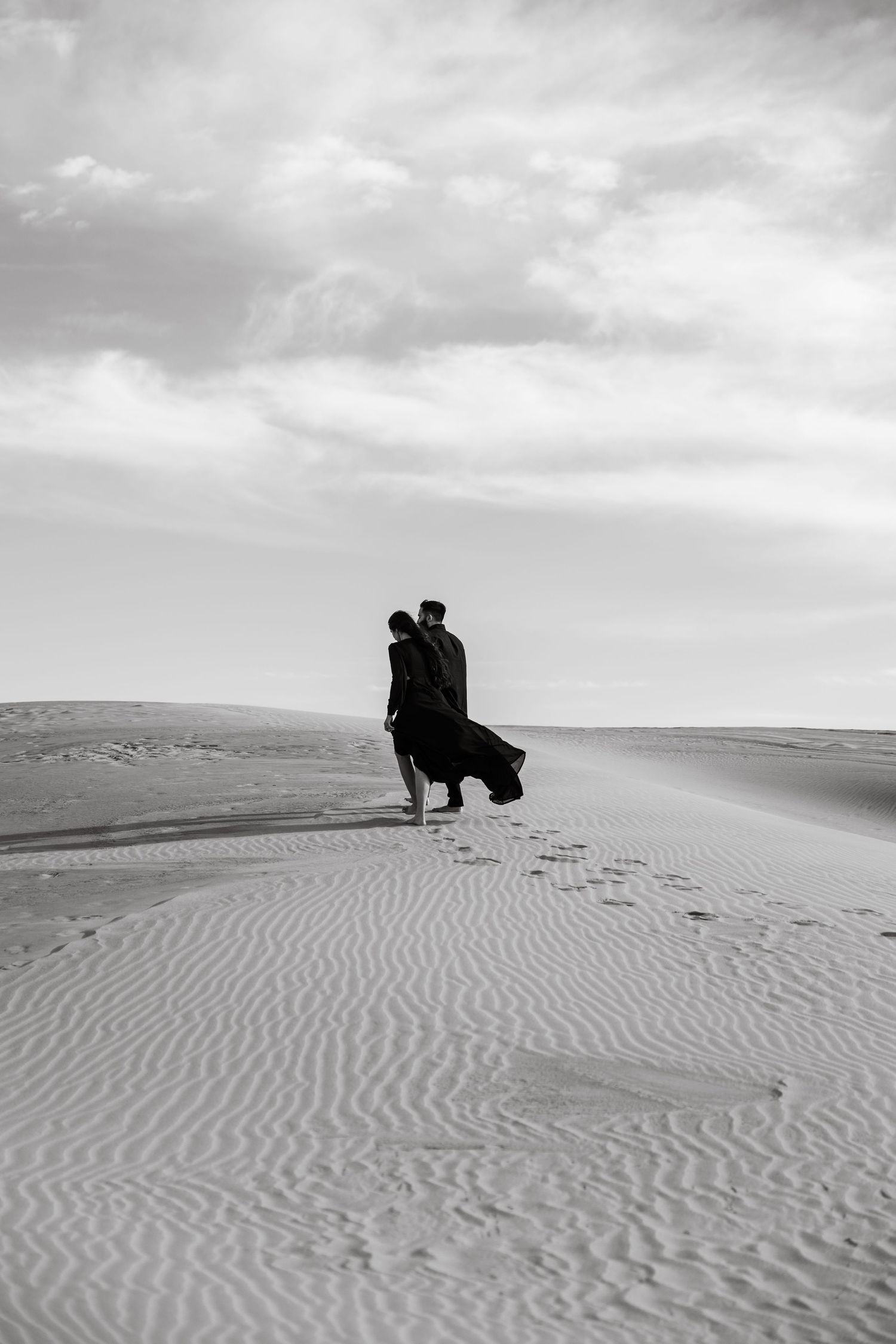 A silhouetted figure stands on rippling sand dunes against a cloudy sky in a dramatic black and white landscape.