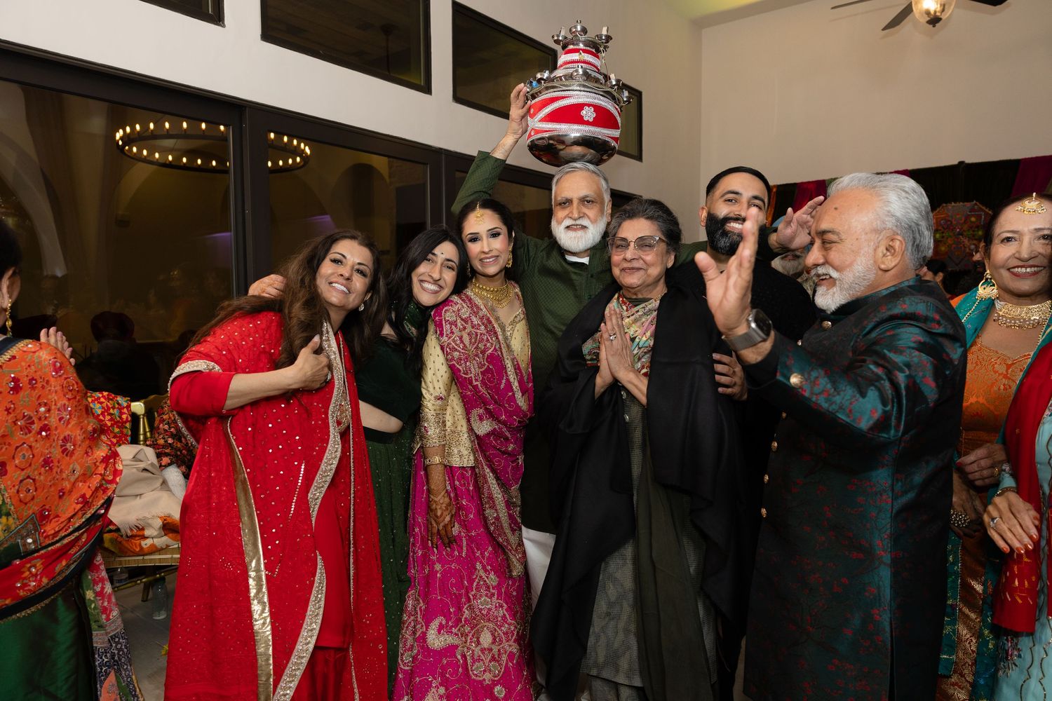 A group celebrates at a festive Indian wedding reception with bright red and pink traditional attire.