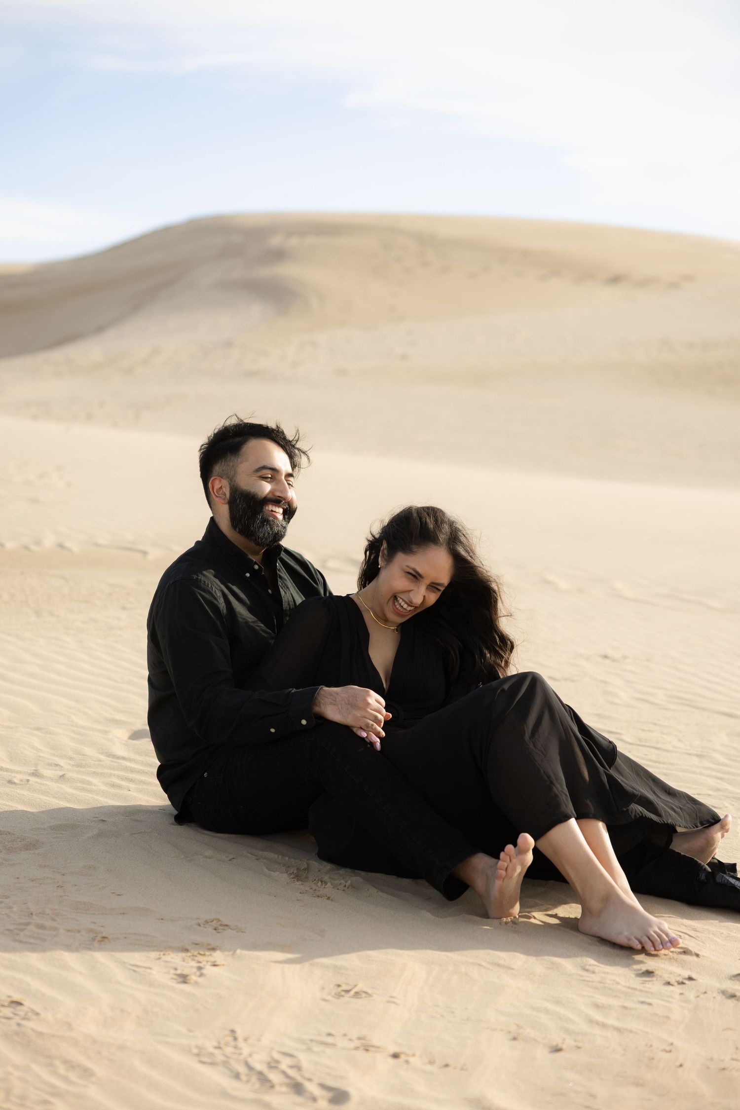 Couple in black clothing sitting together on sandy dunes in a desert landscape.