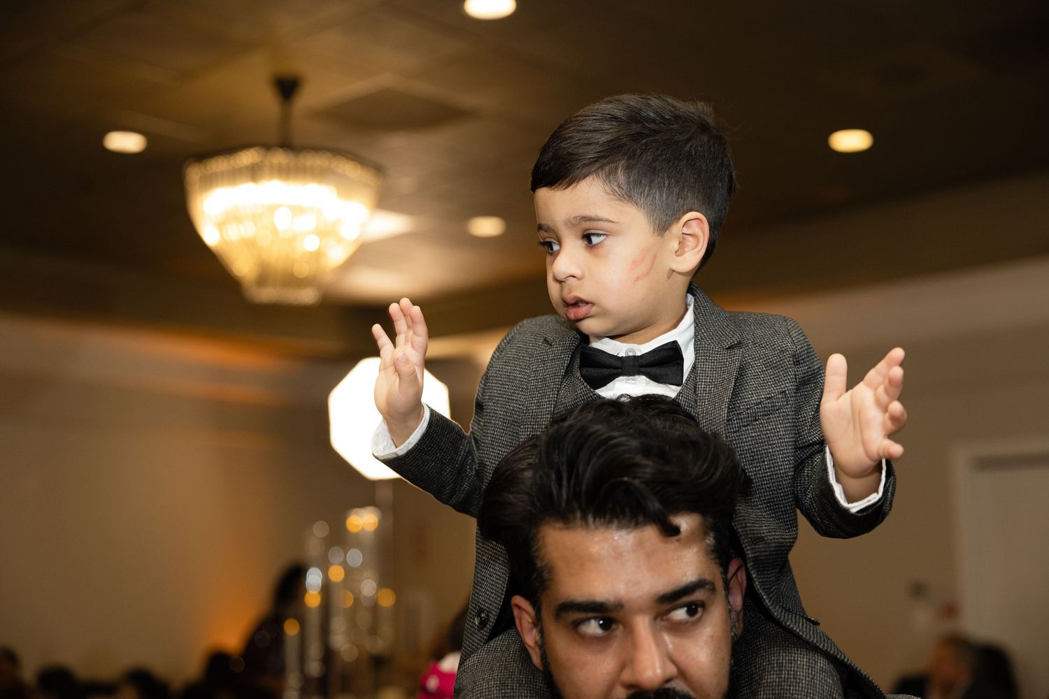 A person in formal attire performs an energetic dance move during a wedding reception at an indoor venue.