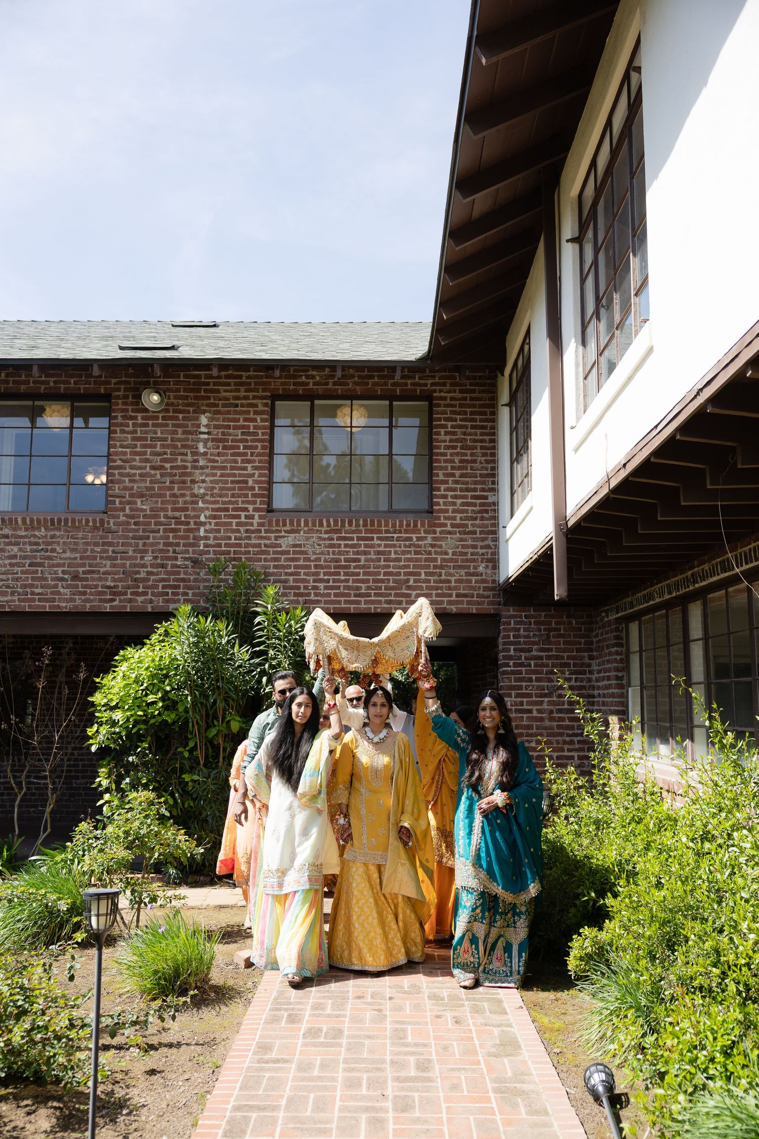 A group in colorful period costumes stands on a brick walkway outside a rustic brick house with overhanging eaves.