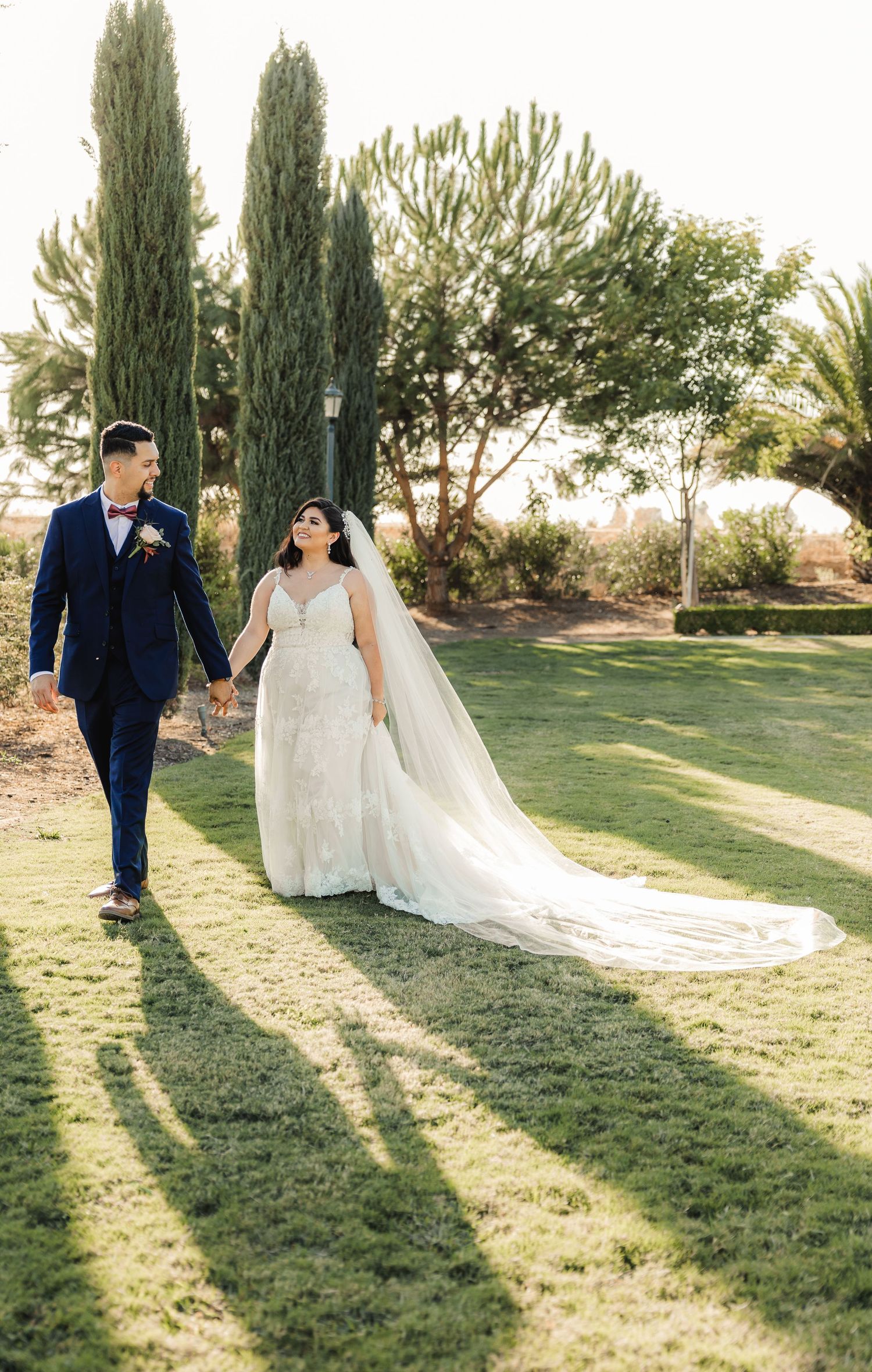 A couple walks hand in hand on manicured lawn with cypress trees and trailing wedding veil.