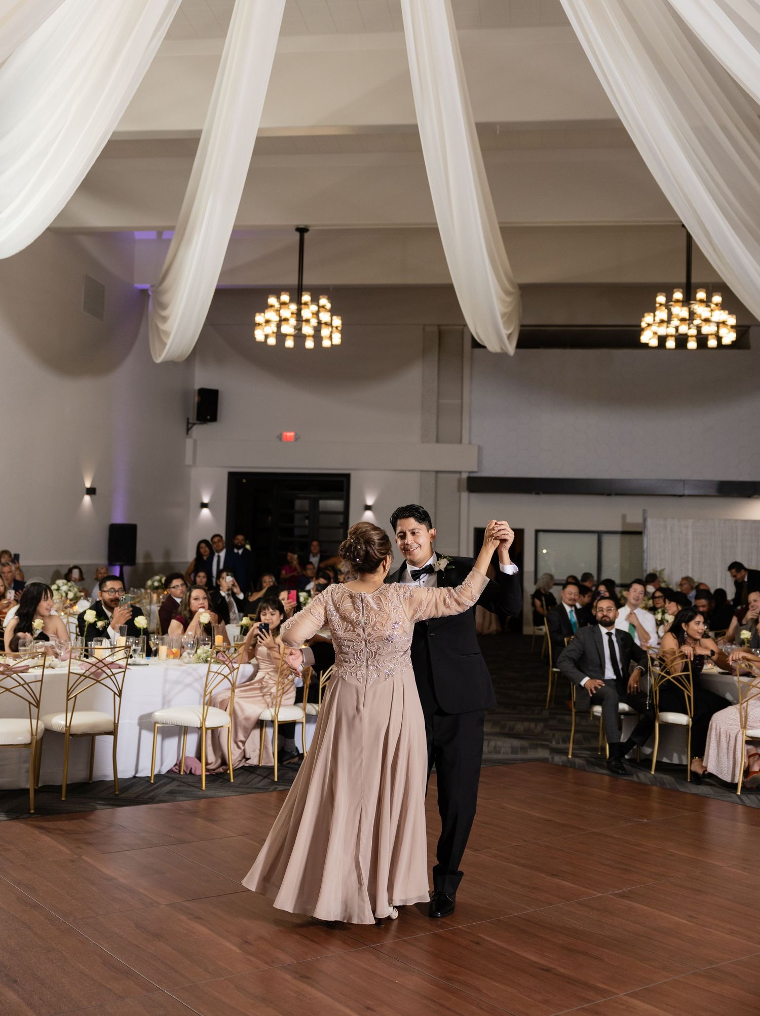 A couple performs their first dance at an elegant wedding reception with draped white ceiling and chandelier lighting.