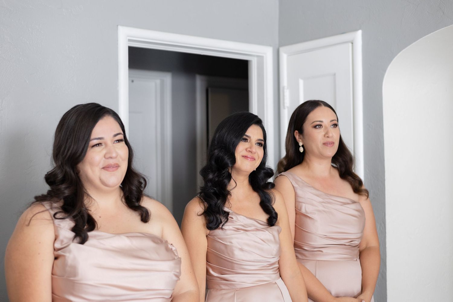 Three bridesmaids wearing matching blush satin dresses pose together in a bright room.