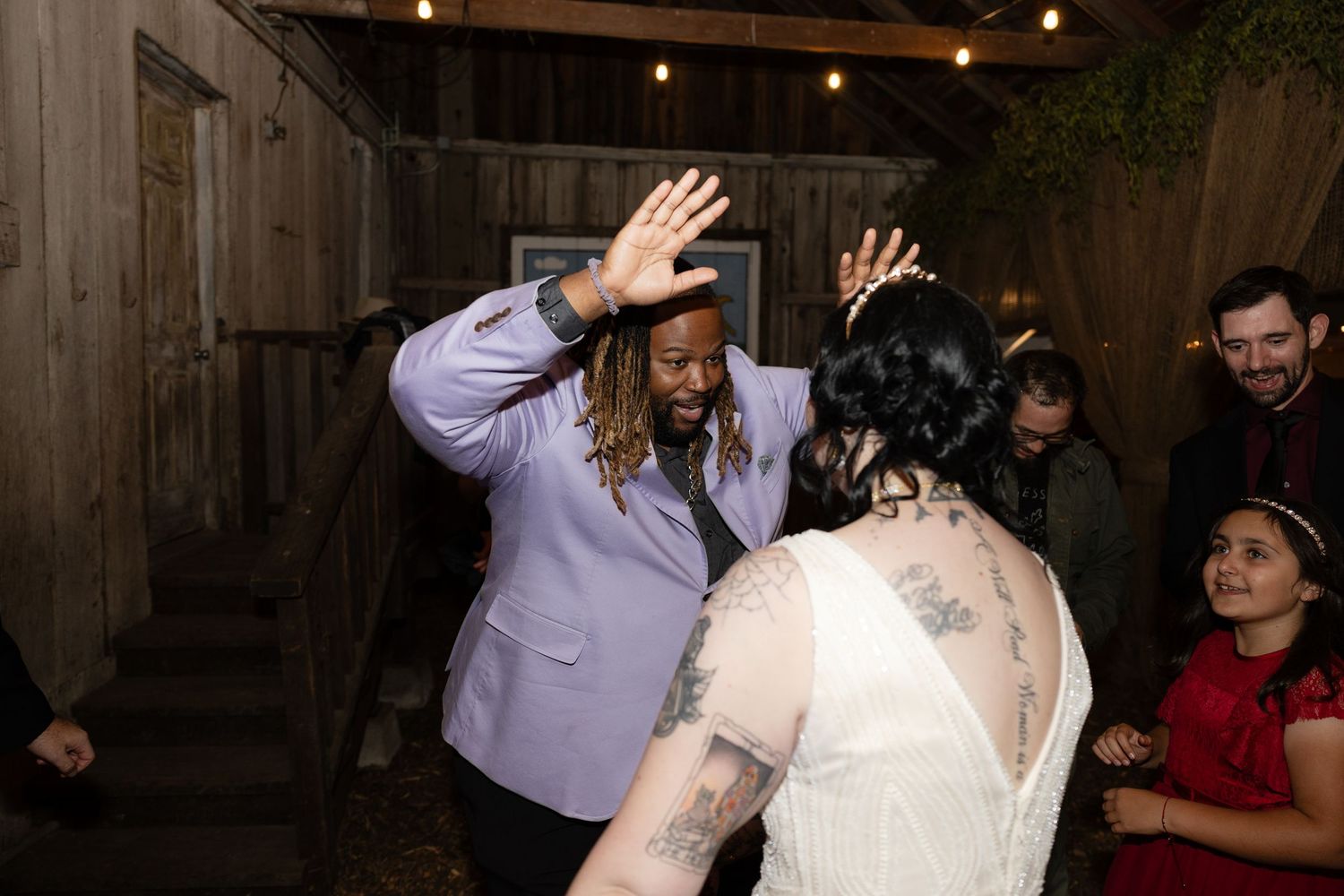 Wedding guests dancing and celebrating beneath string lights in a rustic barn setting.