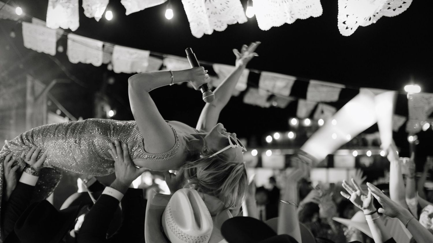 Black and white photo of a crowd-surfing performer at an outdoor concert with string lights and papel picado.