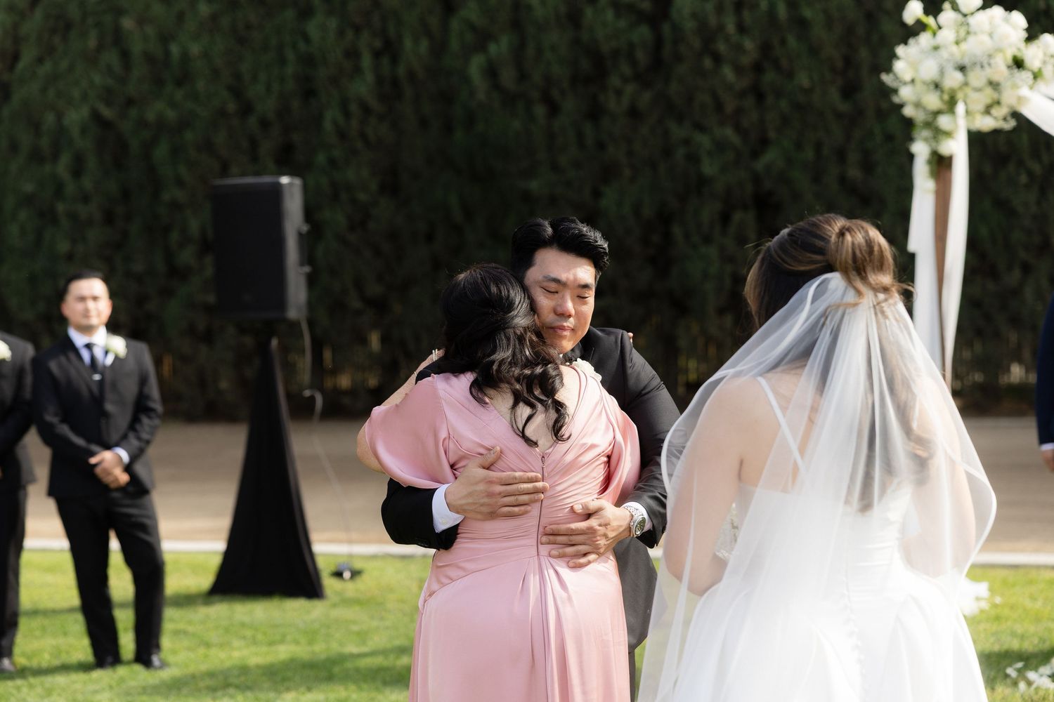 Groom in black suit embraces woman in pink dress as bride in veil stands nearby at outdoor wedding.