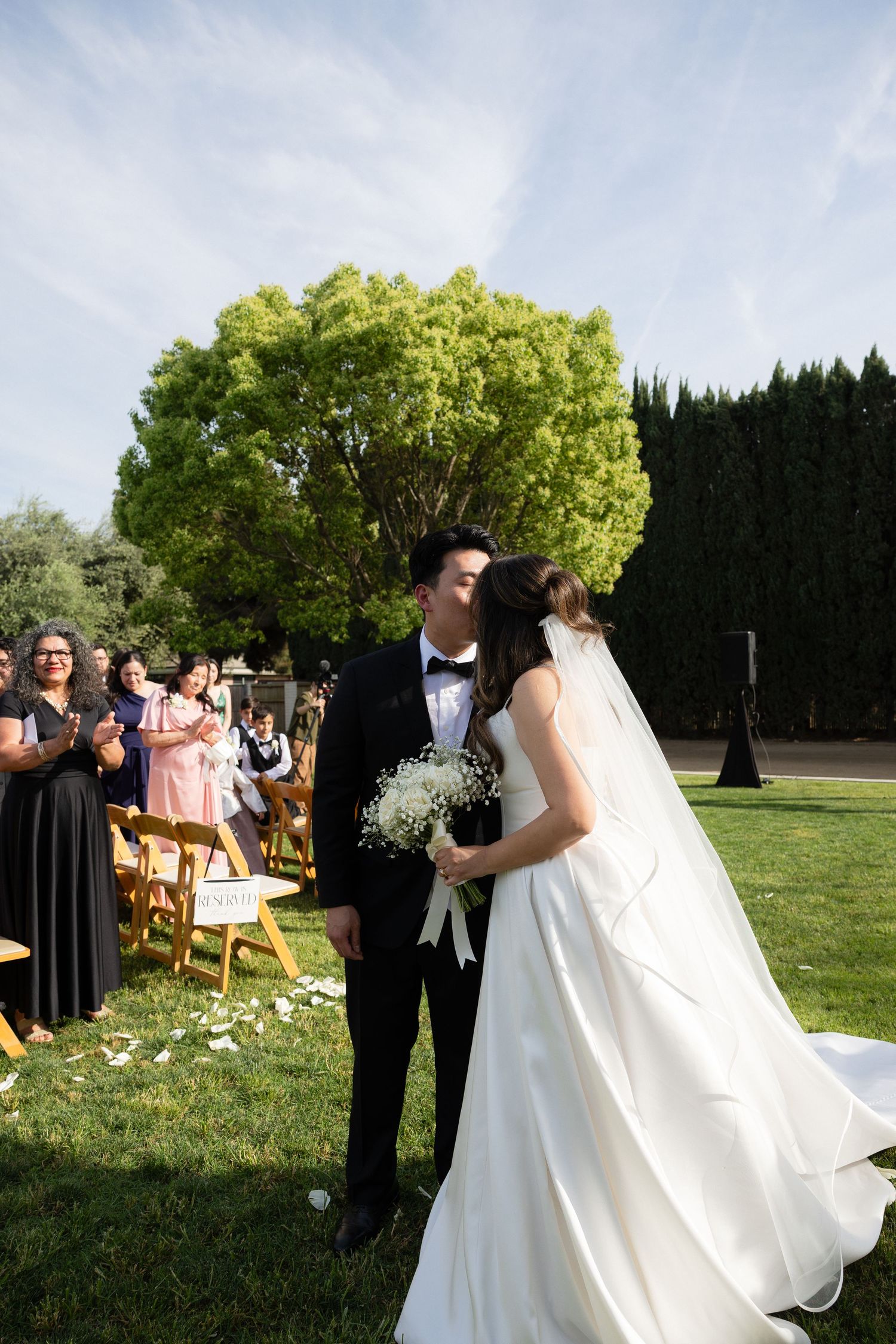 Bride and groom share a first kiss during outdoor wedding ceremony on green lawn with guests applauding.
