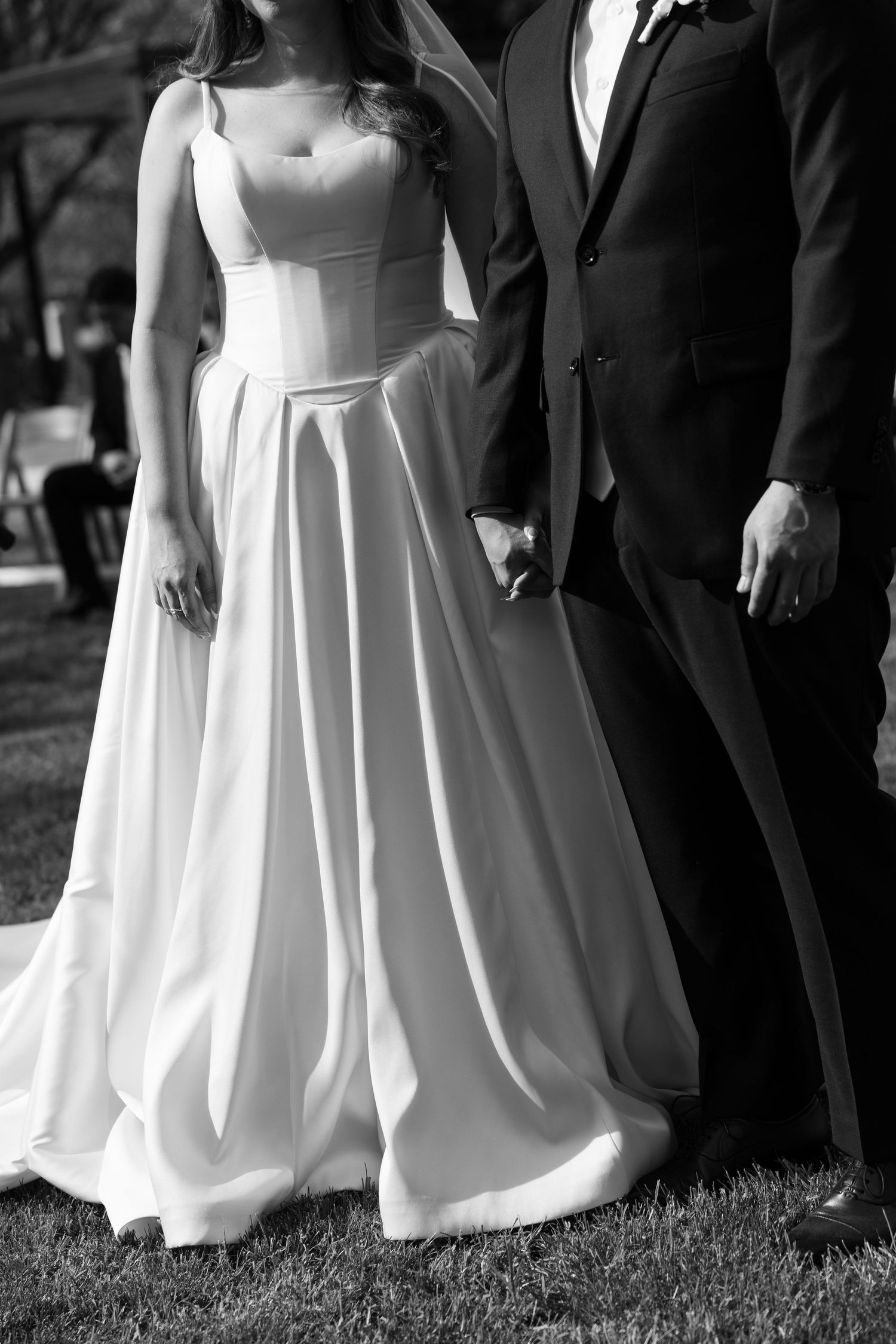 Black and white photo of a bride in elegant A-line gown holding hands with groom in dark suit at outdoor wedding.
