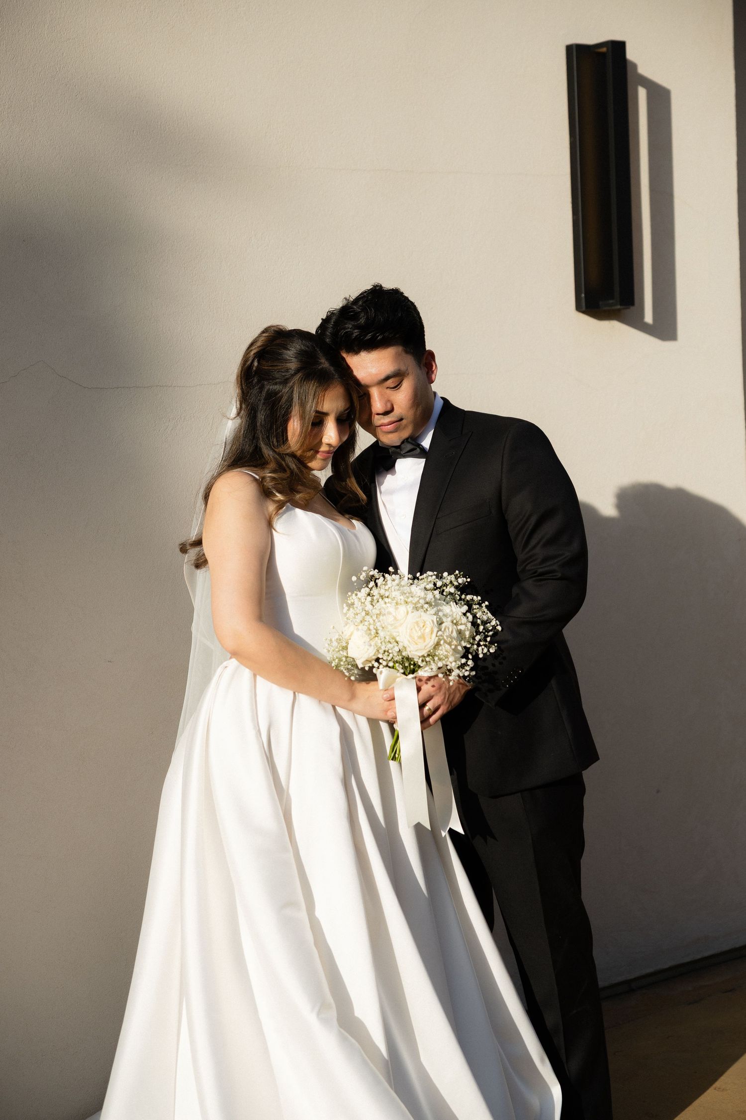 Wedding couple embracing indoors in sunlight, bride in white ball gown with bouquet, groom in black tuxedo with bow tie.