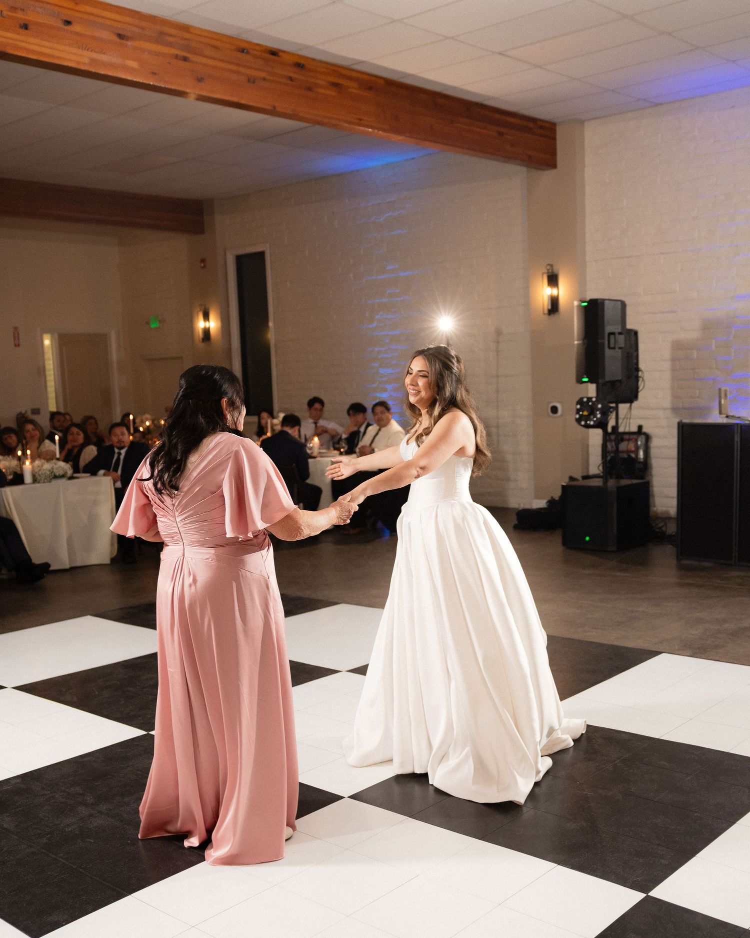 Bride in white gown dancing with bridesmaid in pink dress on checkered dance floor at wedding reception.