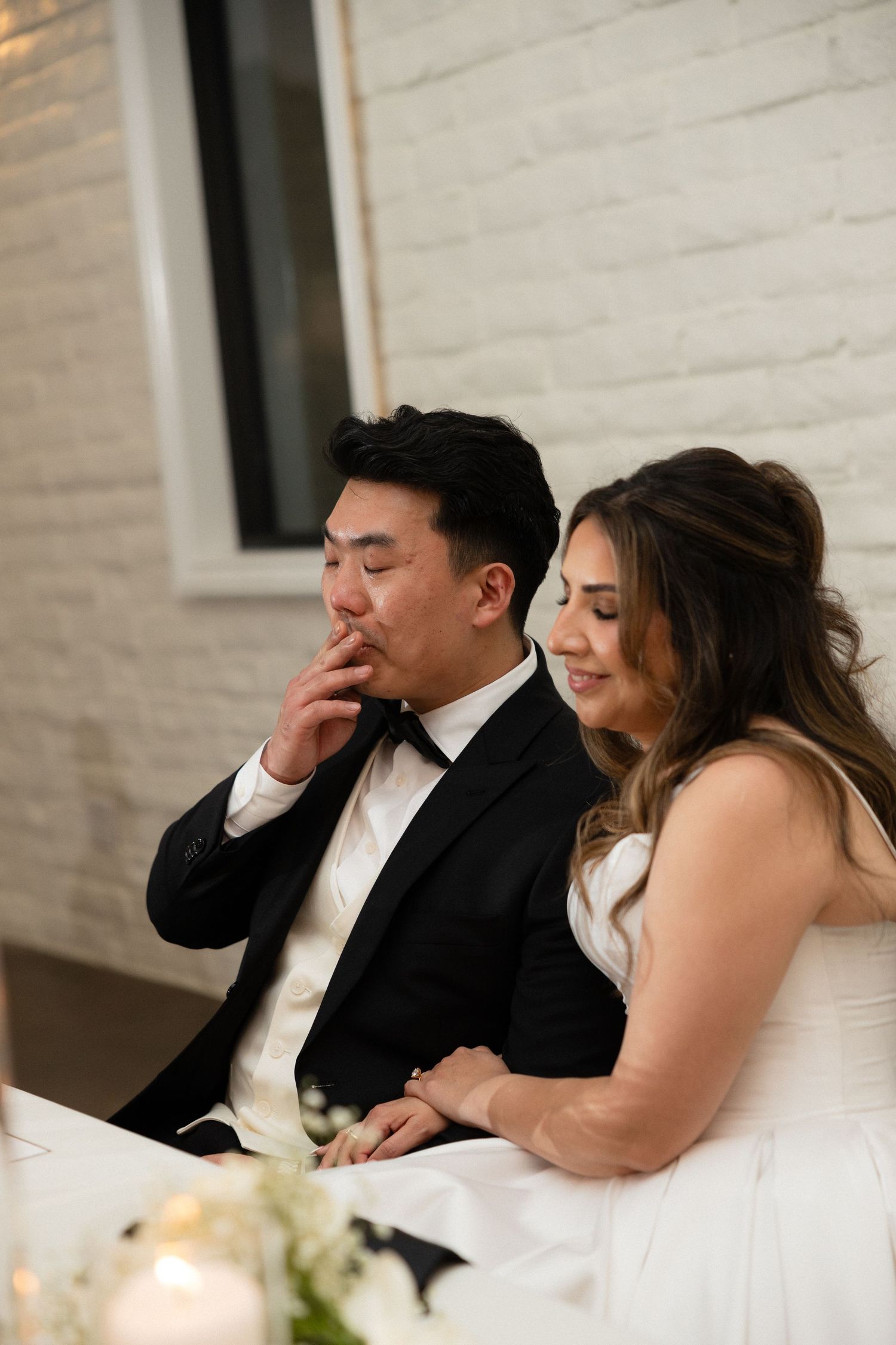 Bride and groom sit together at wedding reception table, groom in black tuxedo, bride in white gown.
