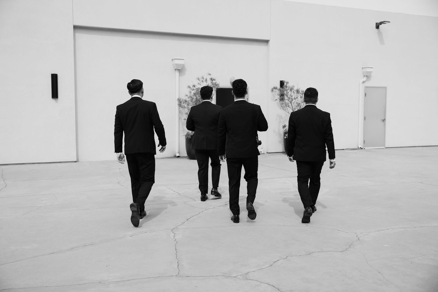 Four groomsmen in black suits walk away toward a bright white wedding venue entrance.