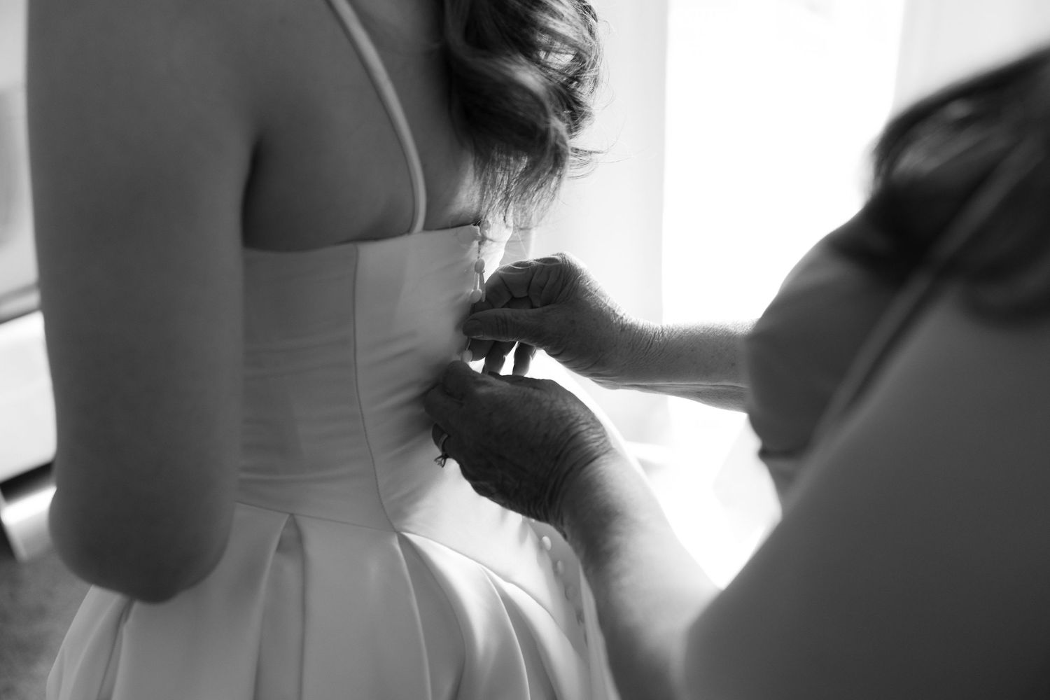Black and white photo of a bride getting her wedding dress buttoned up from behind by a bridesmaid.
