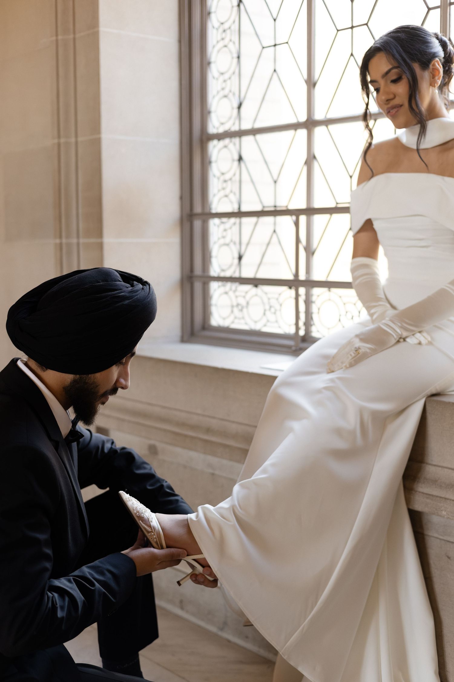 Elegant wedding photo shoot with couple in white dress and black suit posing by sunlit window.
