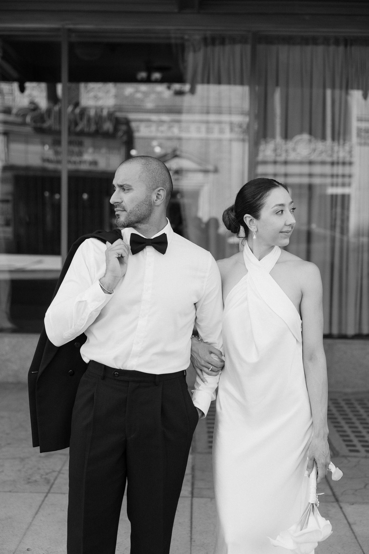 Black and white photo of elegant bride and groom holding hands outside, groom in tuxedo with bow tie, bride in halter wedding gown.