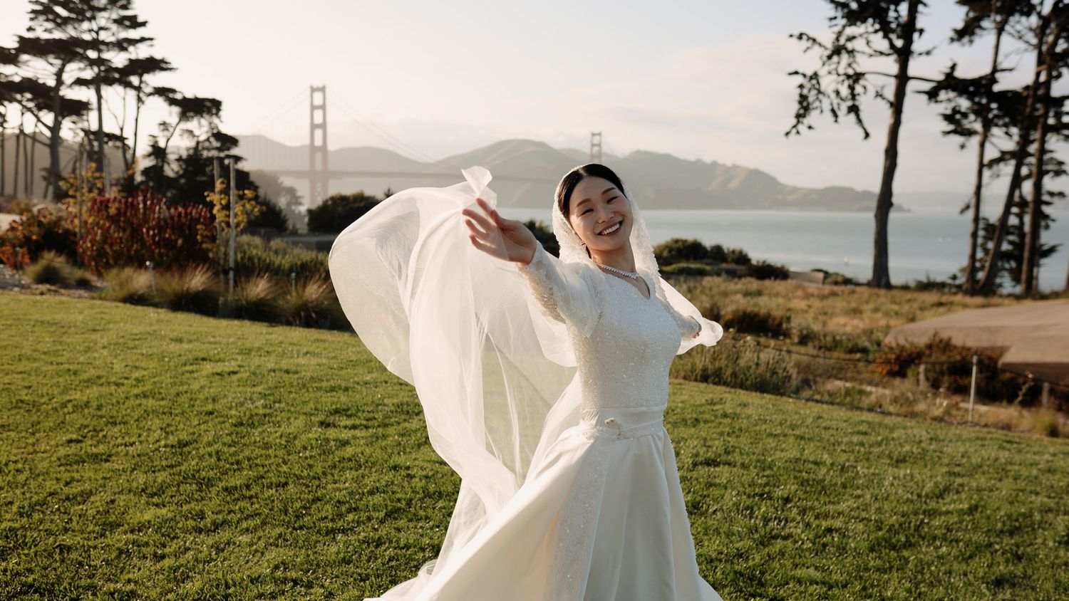 A flowing white wedding dress billows in the wind at a scenic overlook with the Golden Gate Bridge visible in the distance.