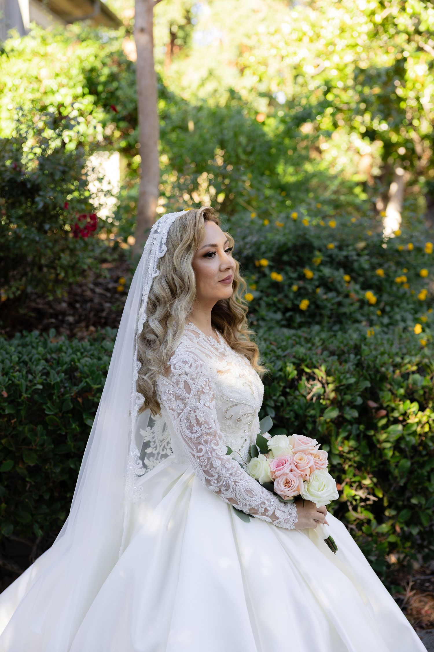 Bride in long-sleeve lace gown holding pink roses against garden backdrop.