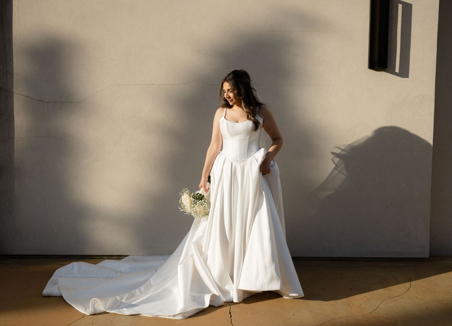 Bride in elegant white ball gown holding bouquet, standing against sunlit wall.
