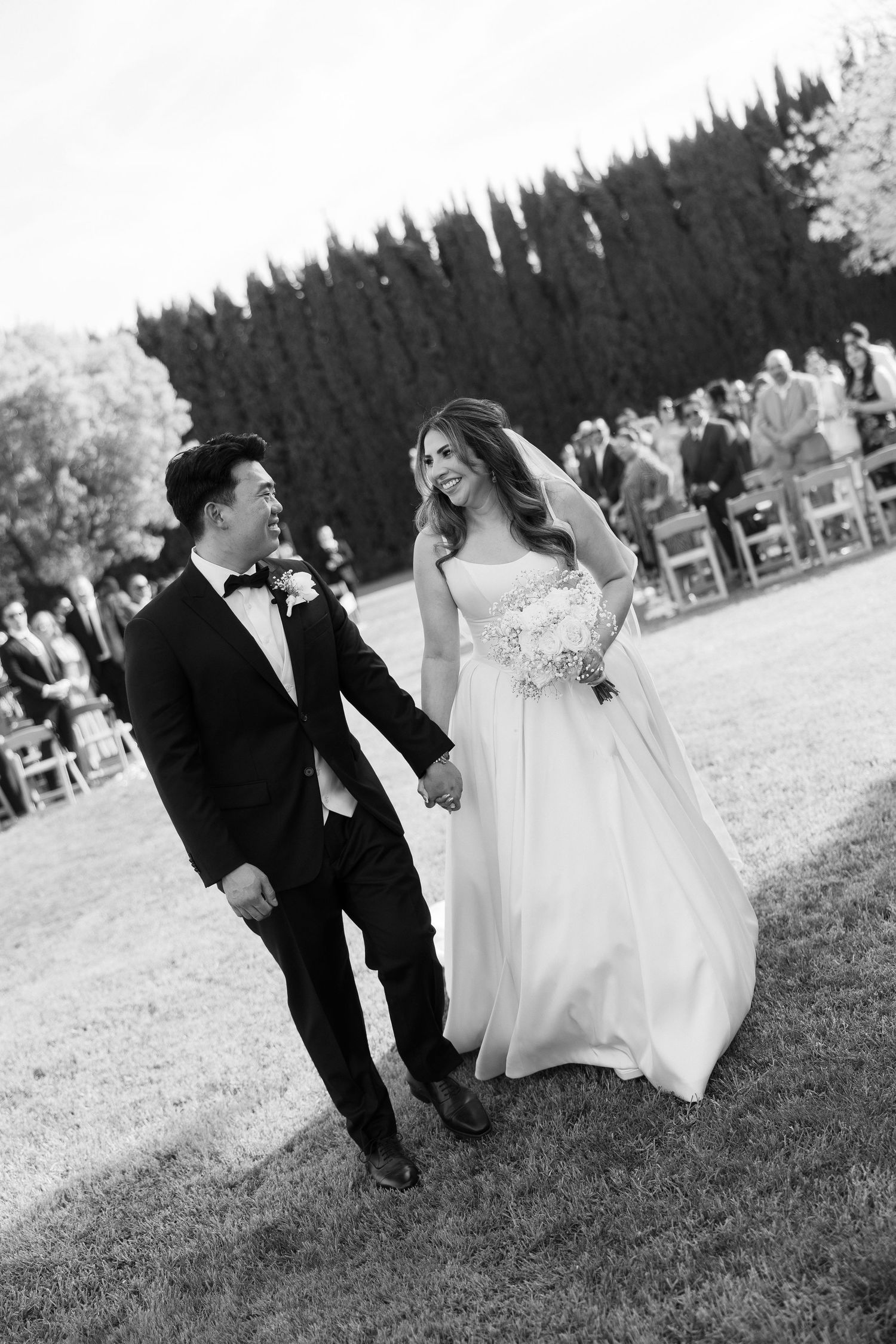Newlyweds walk hand in hand down the aisle on green lawn in black and white photo.