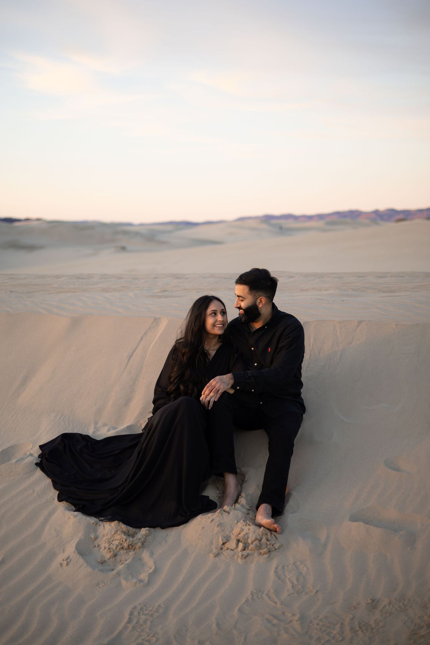 A couple relaxes together on the rippled sand of a vast desert landscape at dusk.