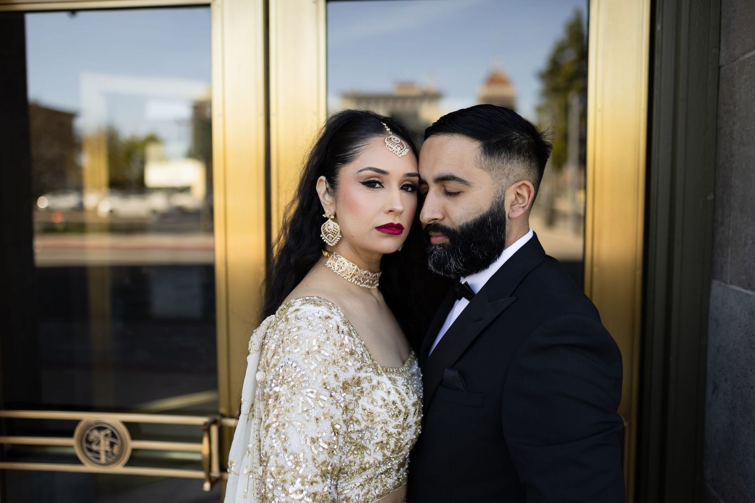 Elegant couple poses outside gold-framed doors at a formal event in black and gold attire.