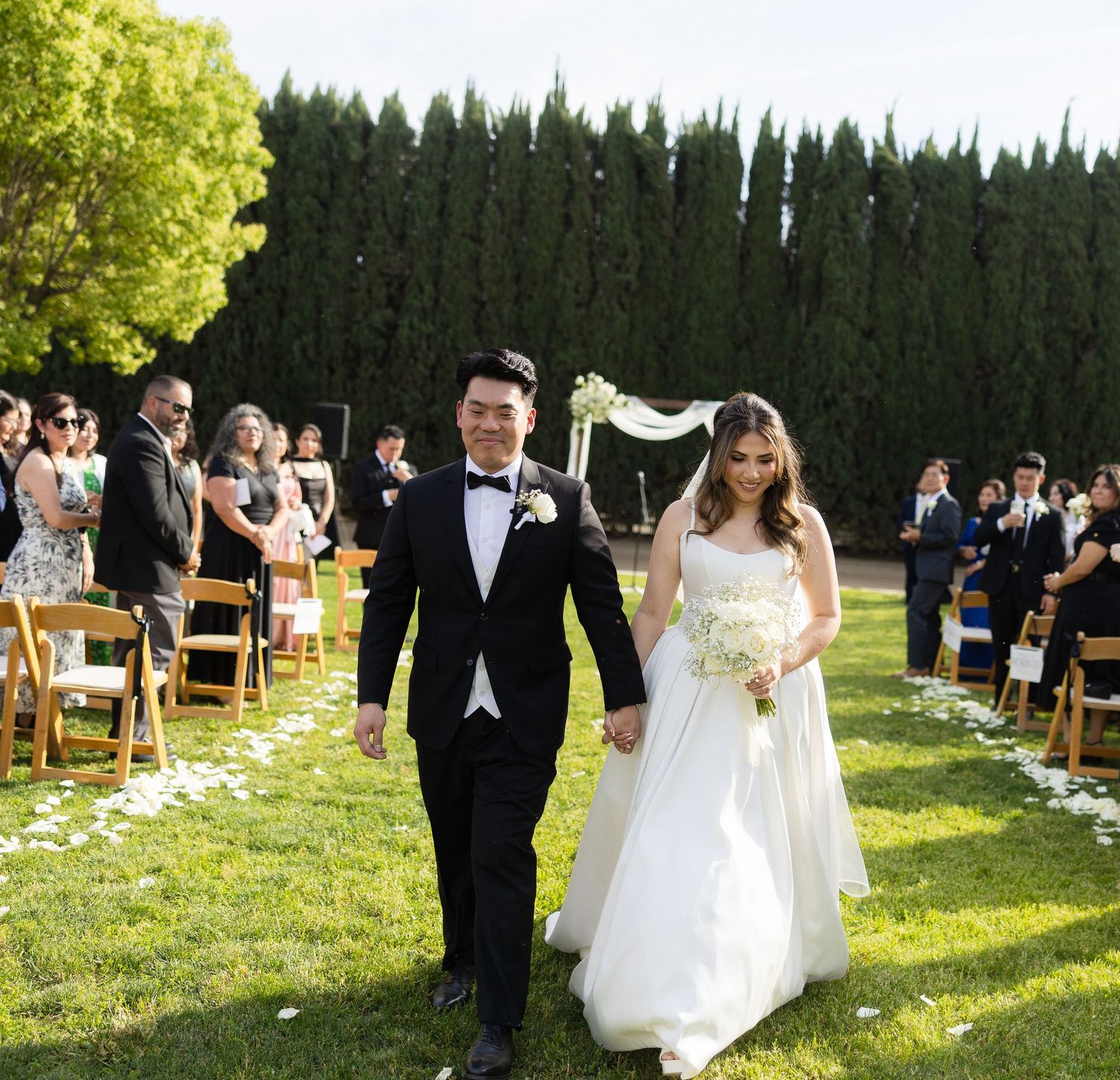 Bride and groom walk down outdoor wedding aisle on green lawn, flanked by guests and tall cypress trees.