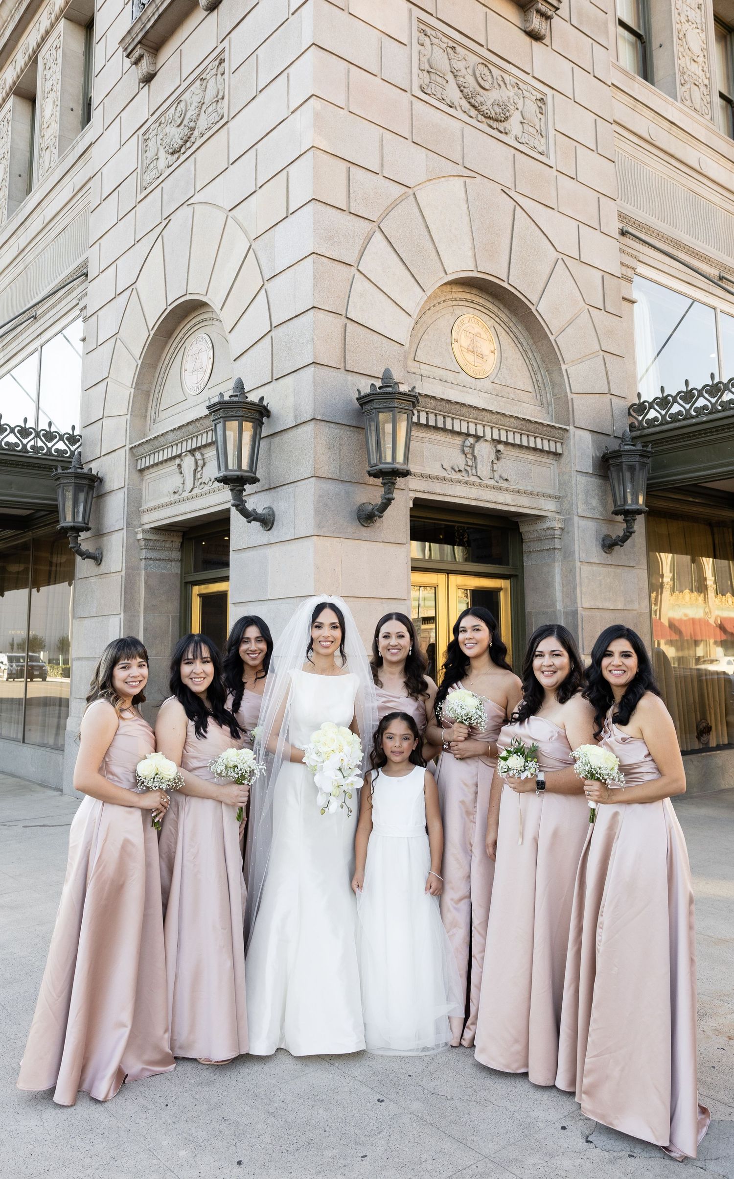 Bridal party in blush pink gowns pose for formal photo outside historic building entrance.