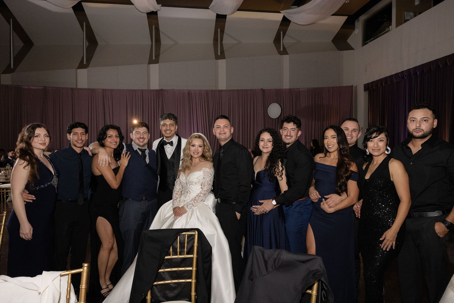Wedding party poses together in formal black attire with bride in white gown at elegant indoor reception venue.