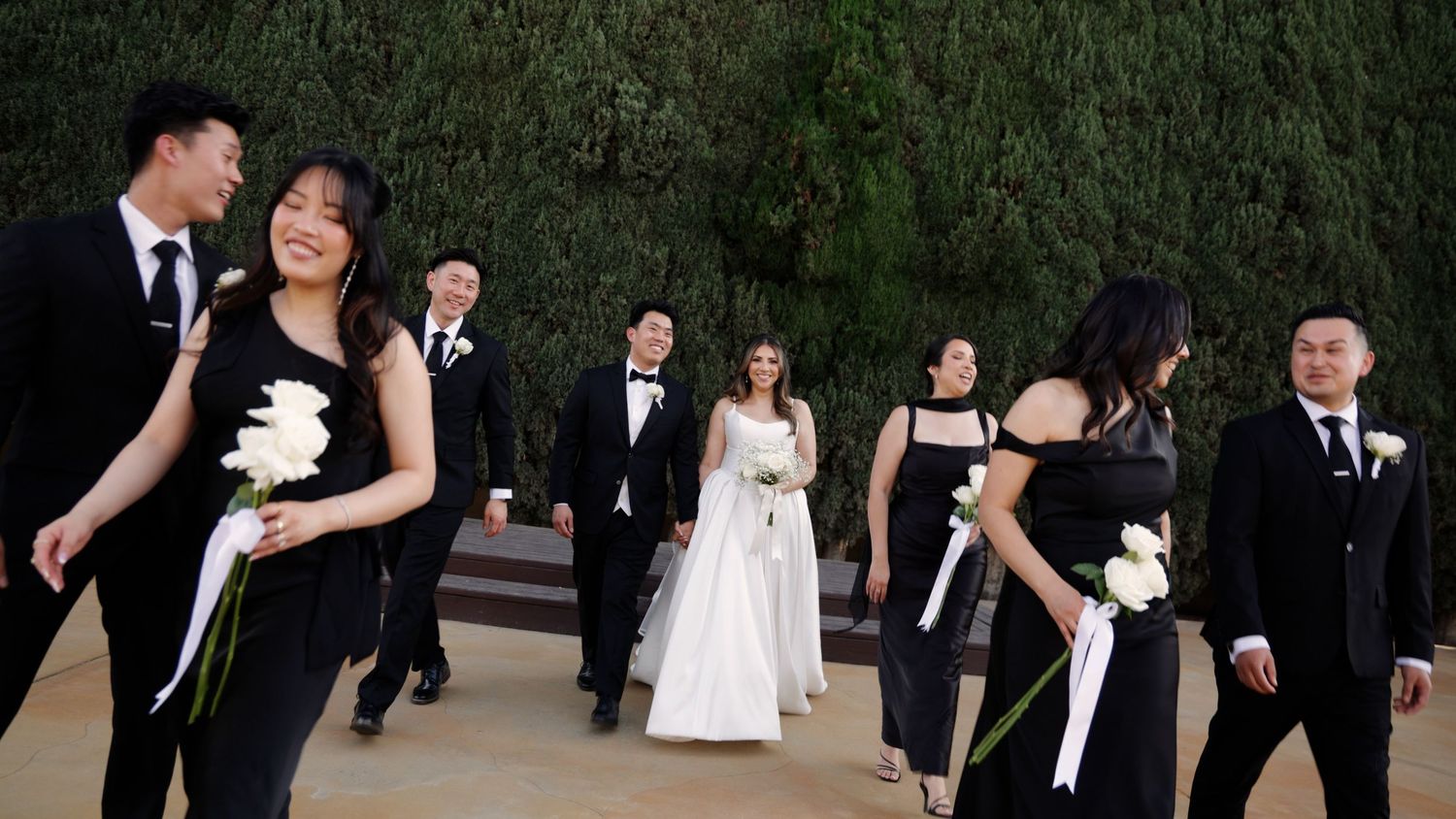 Wedding party in black attire walks outdoors, bride in white gown holding white flowers, greenery wall backdrop.