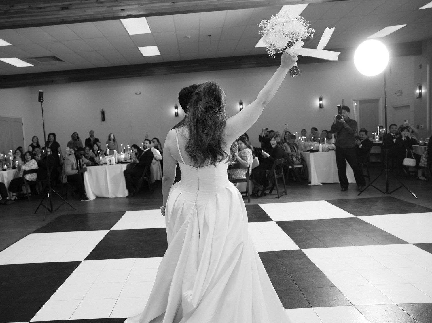 Black and white photo of bride in elegant ball gown raising bouquet triumphantly on checkered dance floor at wedding reception.